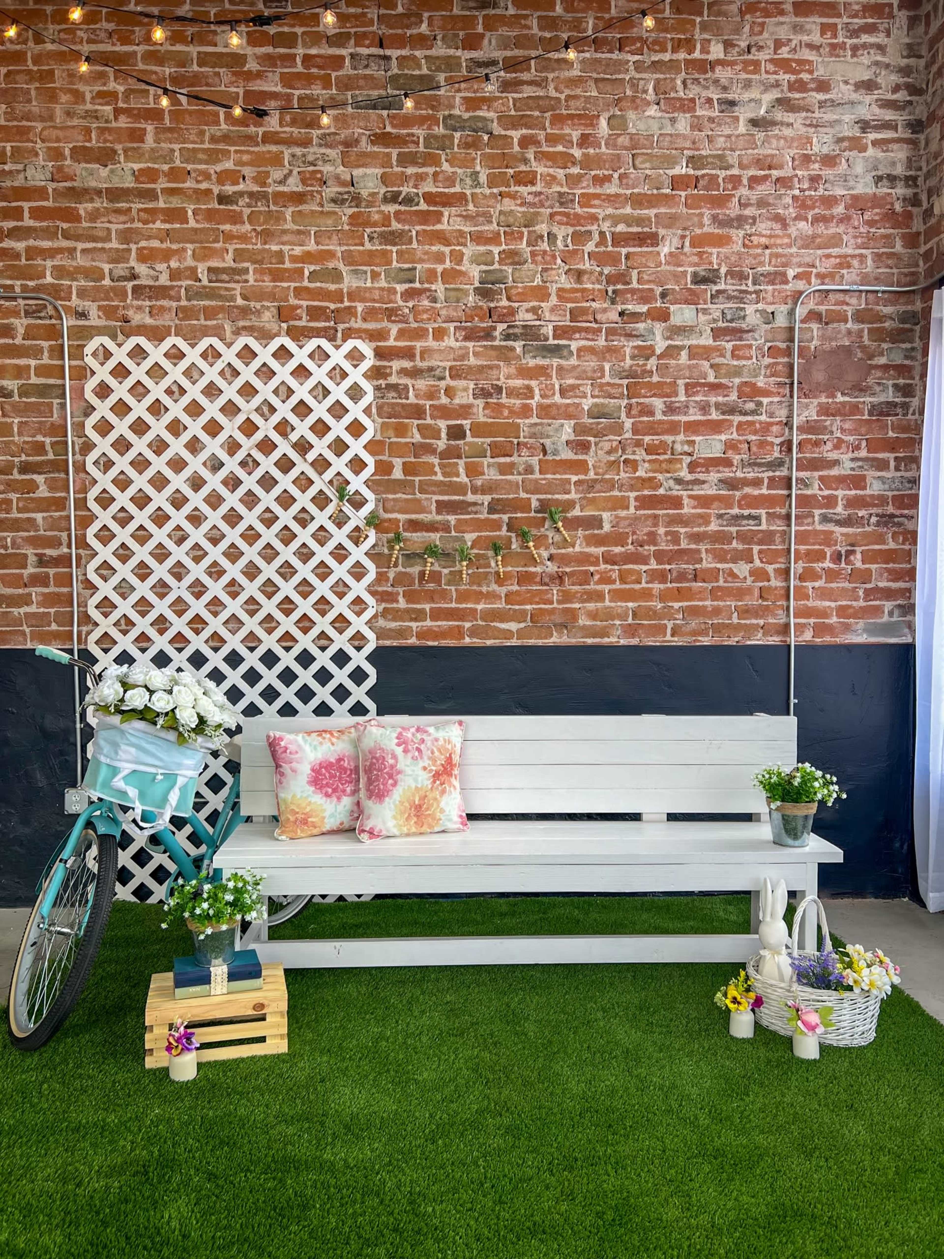 The scene features a white bench with colorful pillows, a bicycle with flowers, and potted plants, set against a brick wall and artificial grass.