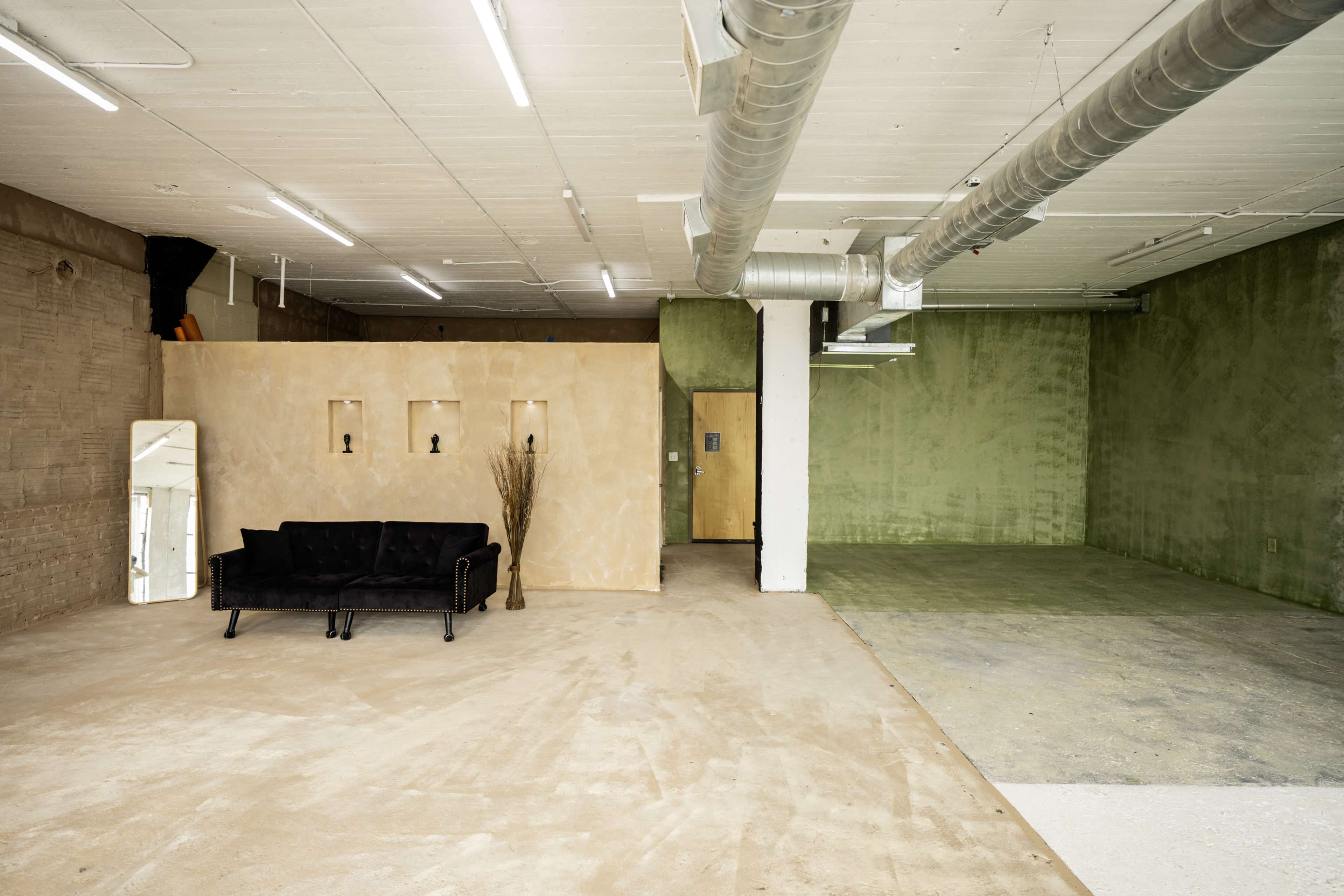 The image shows an empty industrial-style space with a black sofa and a textured peach-colored wall opposite a green wall.