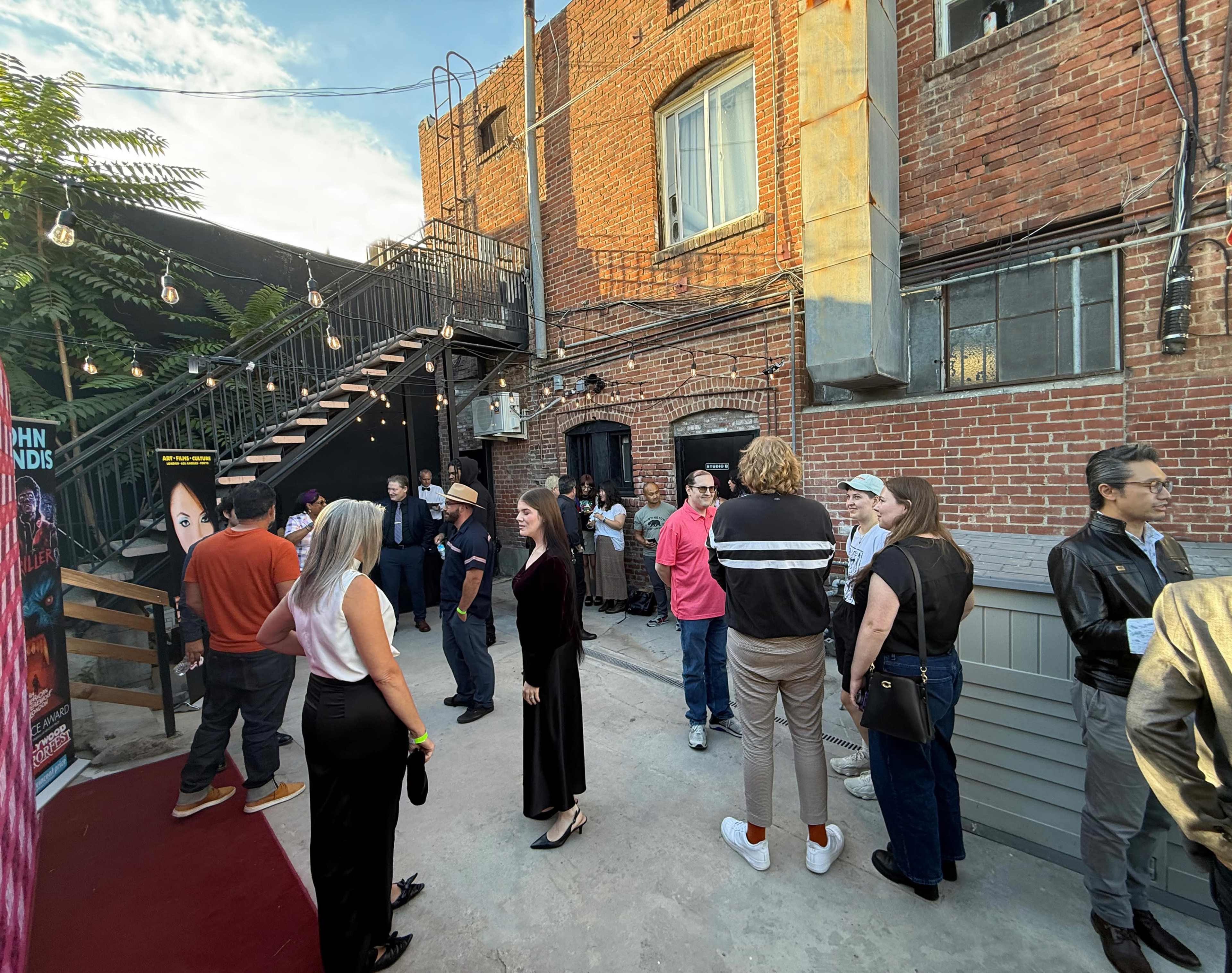 A crowd gathers outside a brick building with a staircase, engaged in conversation under string lights.