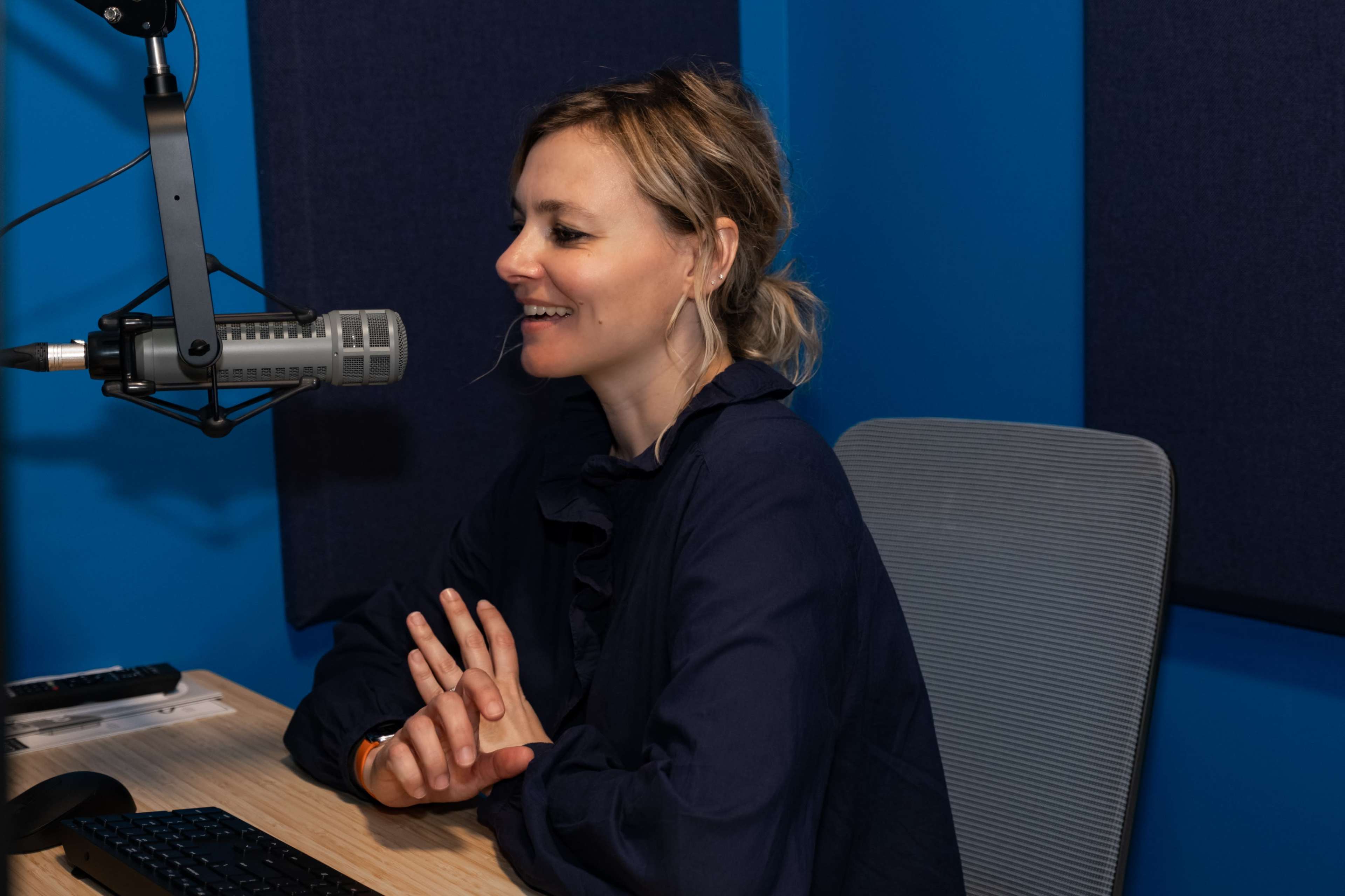 A woman sits at a desk in a recording studio, speaking into a microphone.