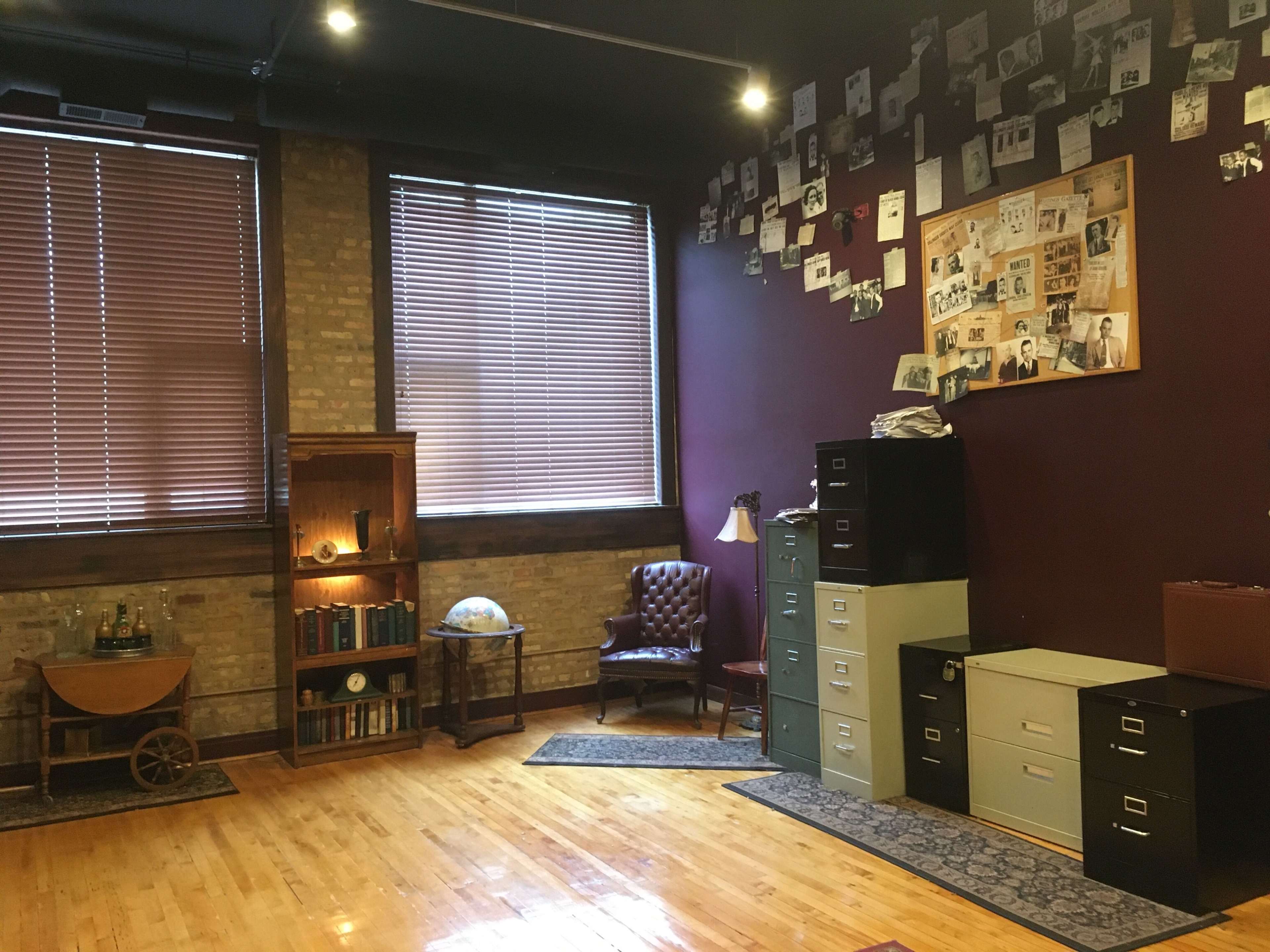 The room features wooden flooring, a large window with blinds, a wall covered in black-and-white photographs, and an arrangement of furniture including a leather chair, globe, bookshelf, and filing cabinets.