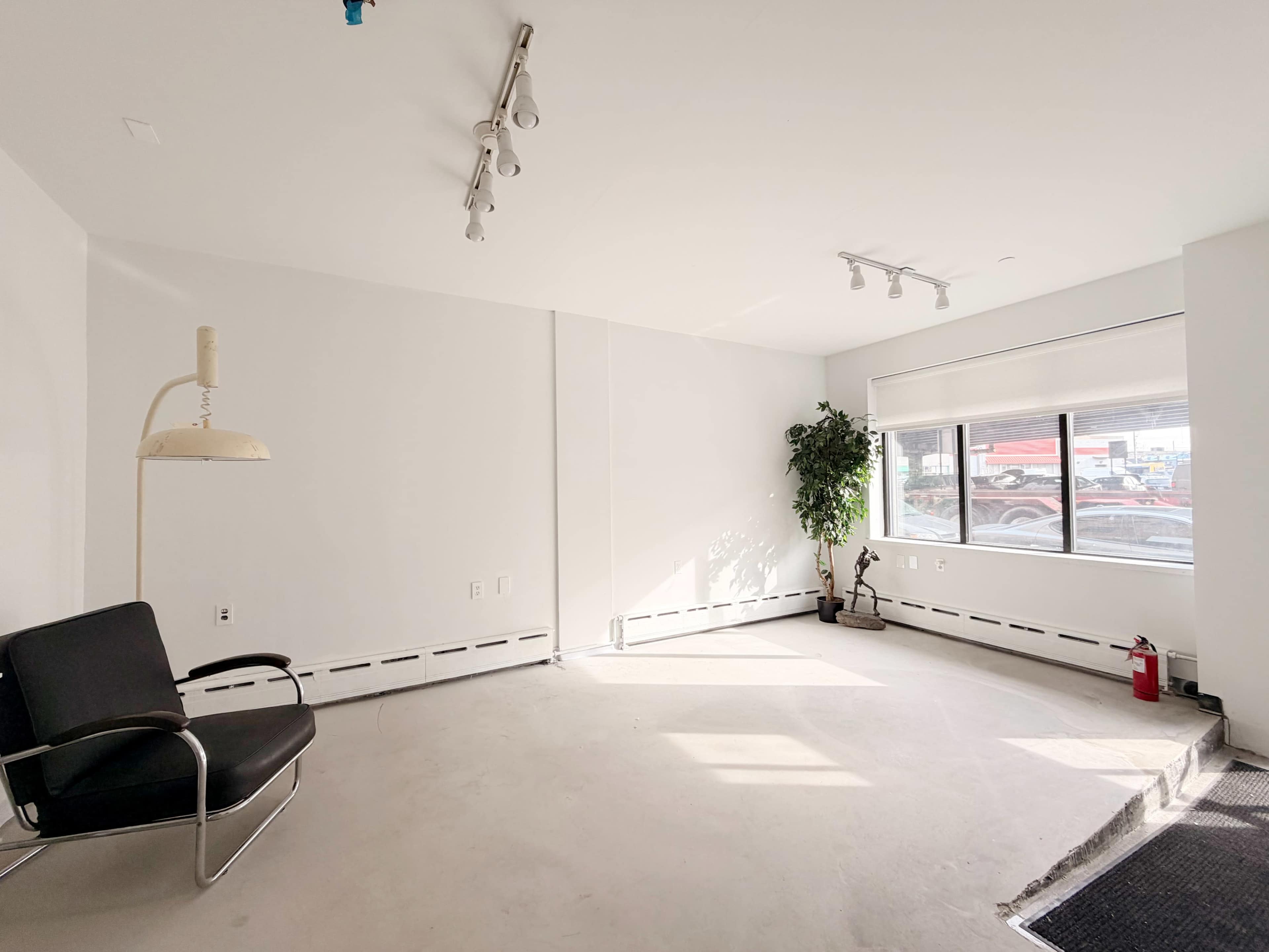 The image shows an empty room with a black chair, a potted plant, a window, and exposed floor.