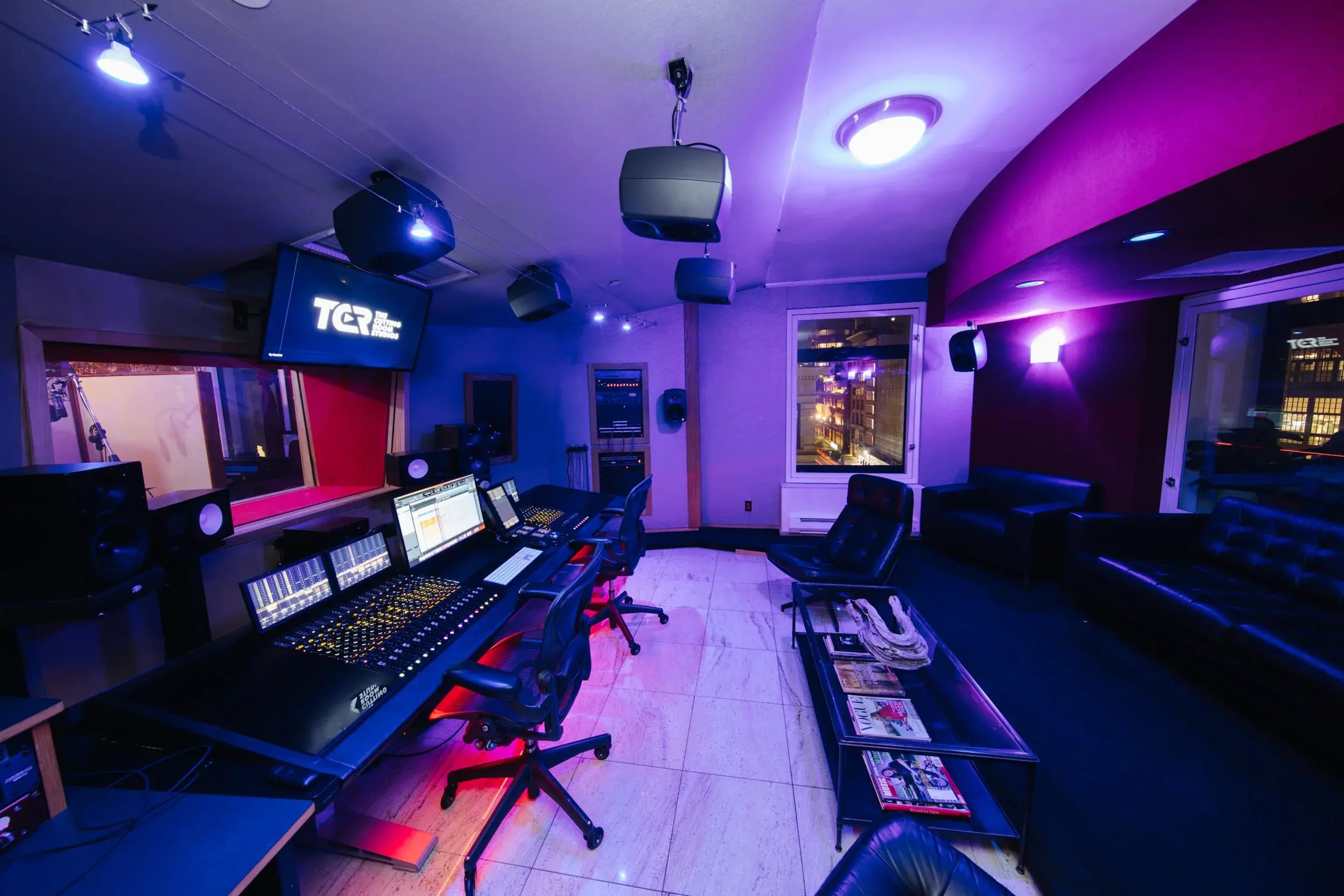 The image depicts a well-equipped music studio with a large mixing desk, speakers, and a comfortable seating area.