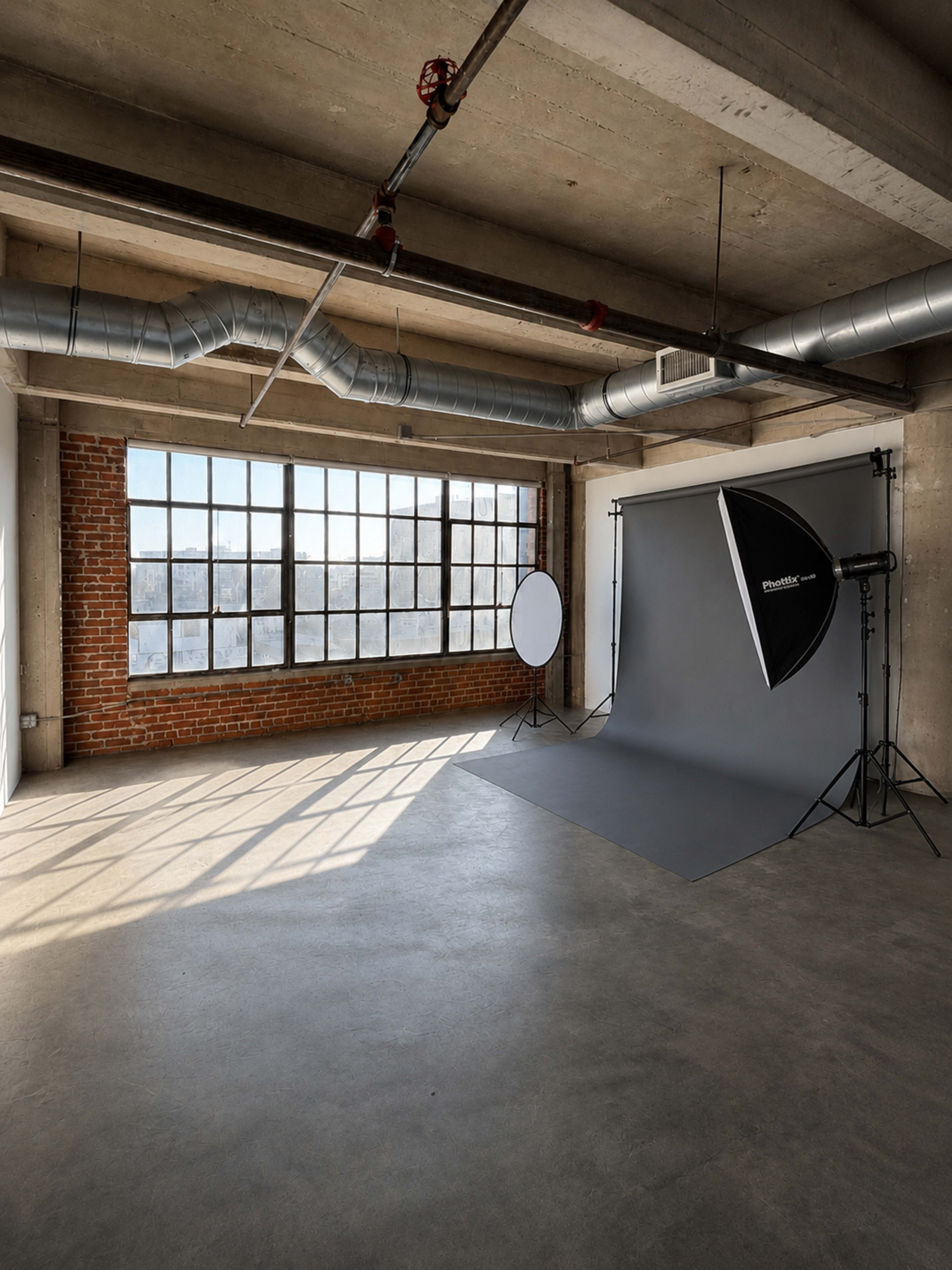 The image shows a spacious photography studio featuring large windows, exposed brick walls, and a gray backdrop set up for a photoshoot.