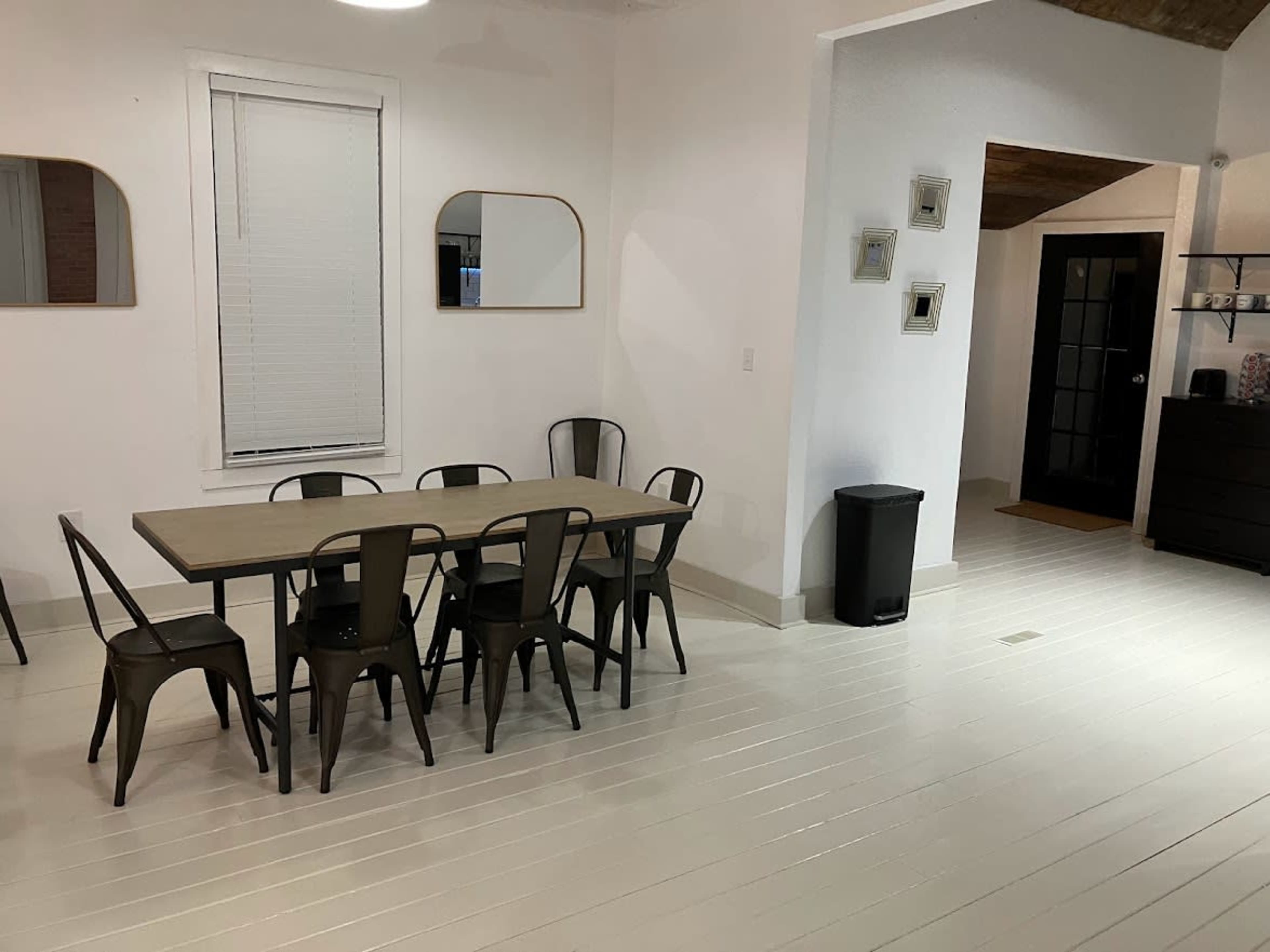 A minimalistic dining area with a long table surrounded by metal chairs and a waste bin in a predominantly white room.