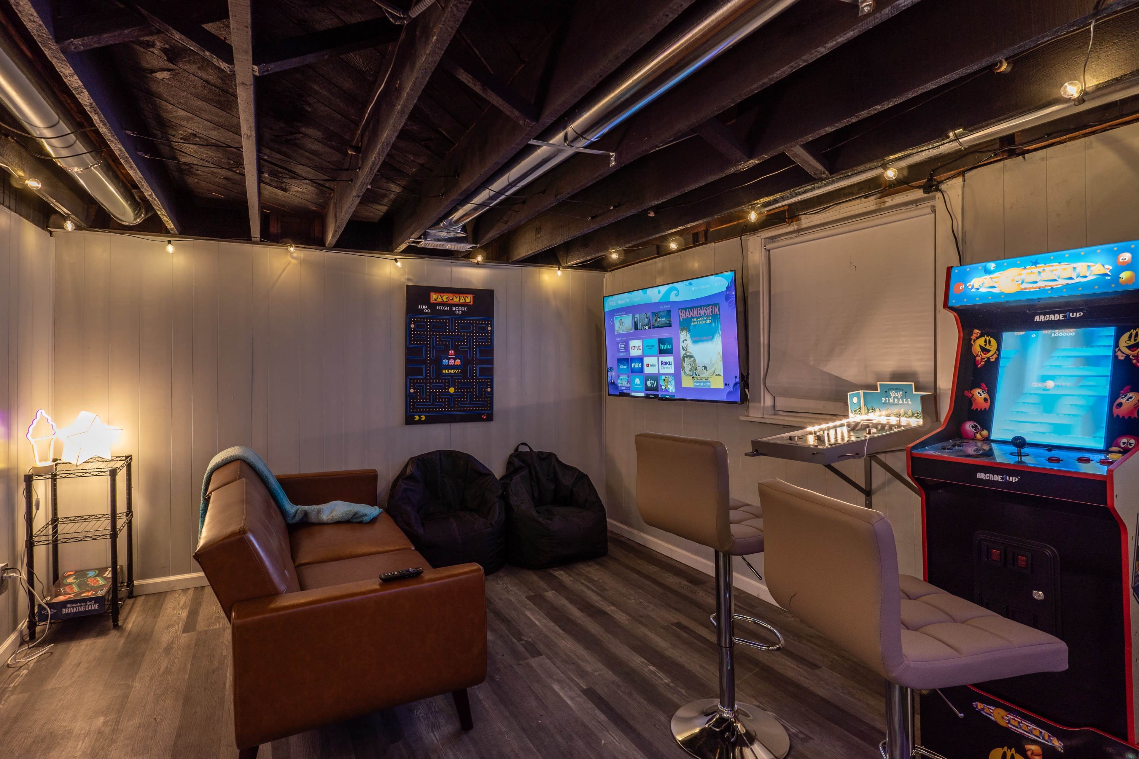 The image depicts a cozy game room featuring a brown couch, bean bag chairs, a bar area with stools, a vintage arcade machine, and two television screens mounted on the wall.