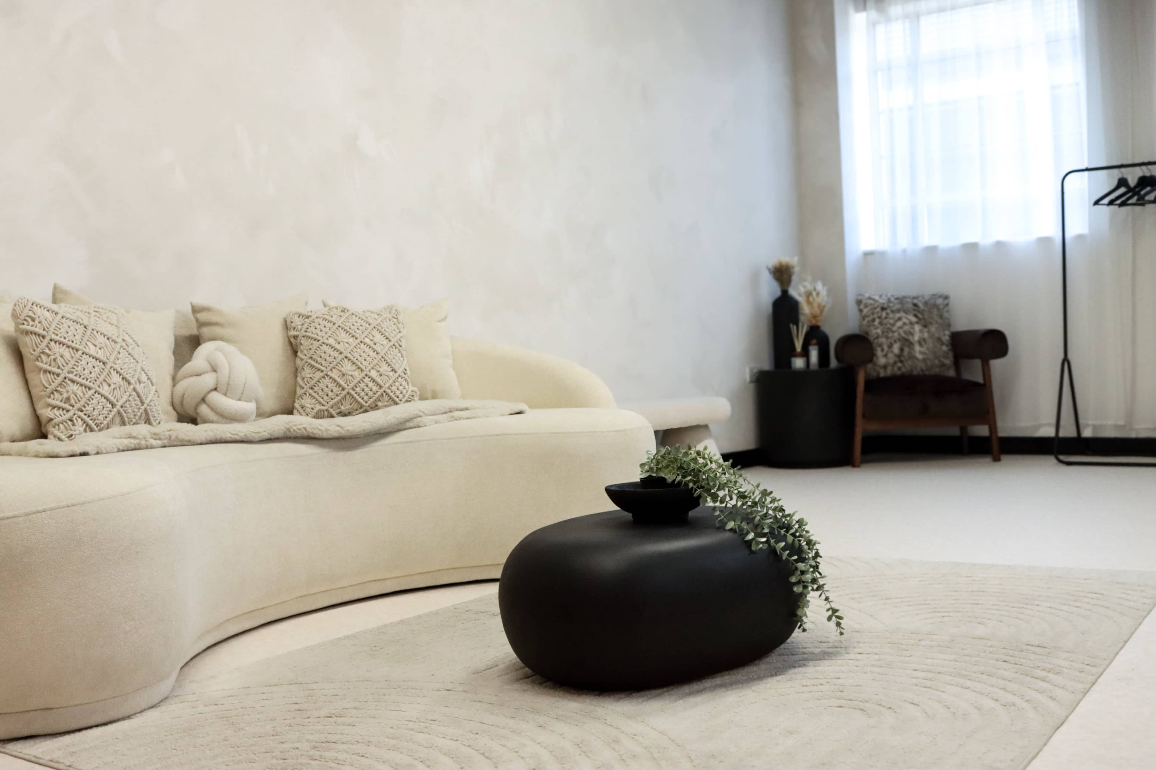 The image features a minimalist living room with a curved beige sofa, a round black coffee table adorned with greenery, and a large window that allows natural light to fill the space.