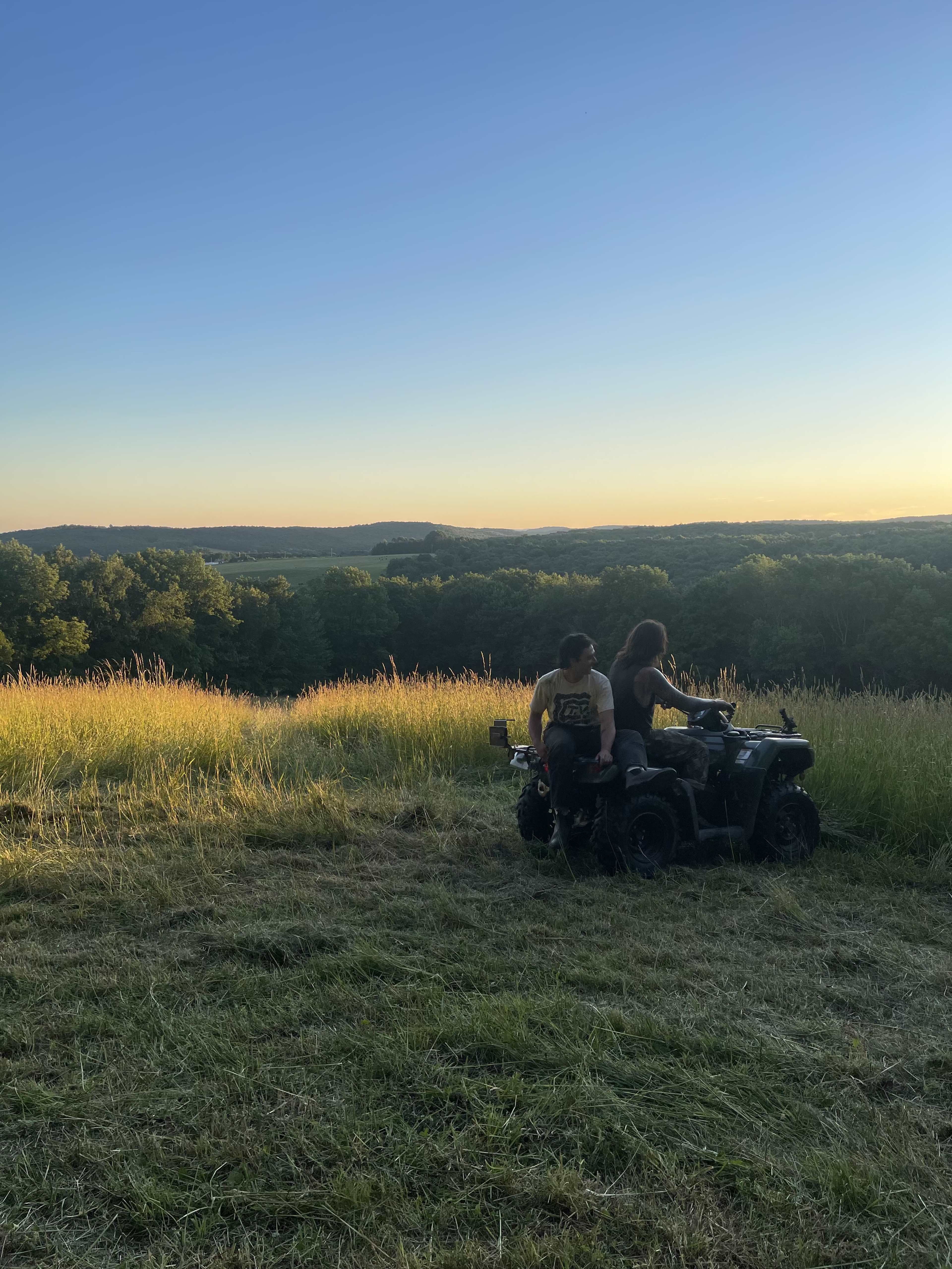 Two people sit on an ATV in a grassy field at sunset, with rolling hills in the background.