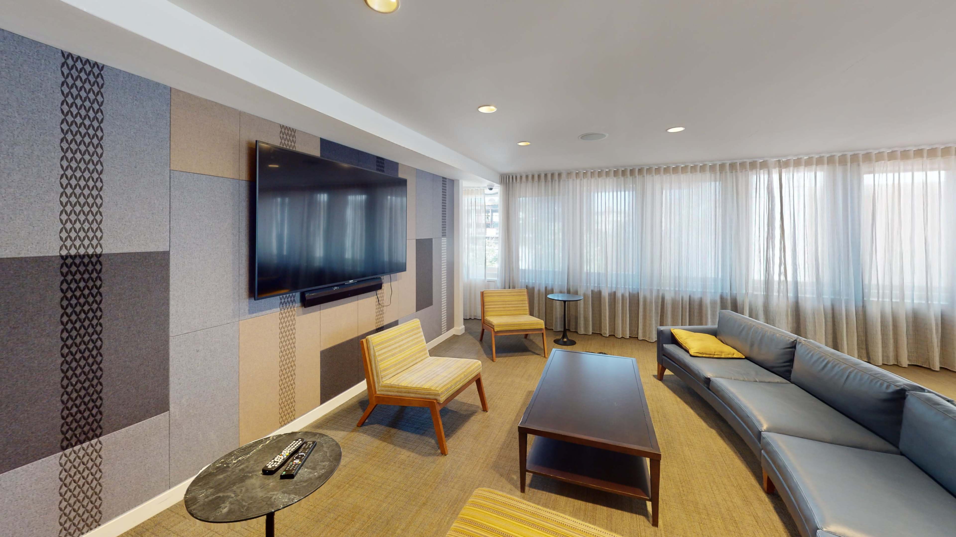 The image shows a modern living room with a large television mounted on the wall, a sectional sofa, two armchairs, a coffee table, and large windows with sheer curtains.