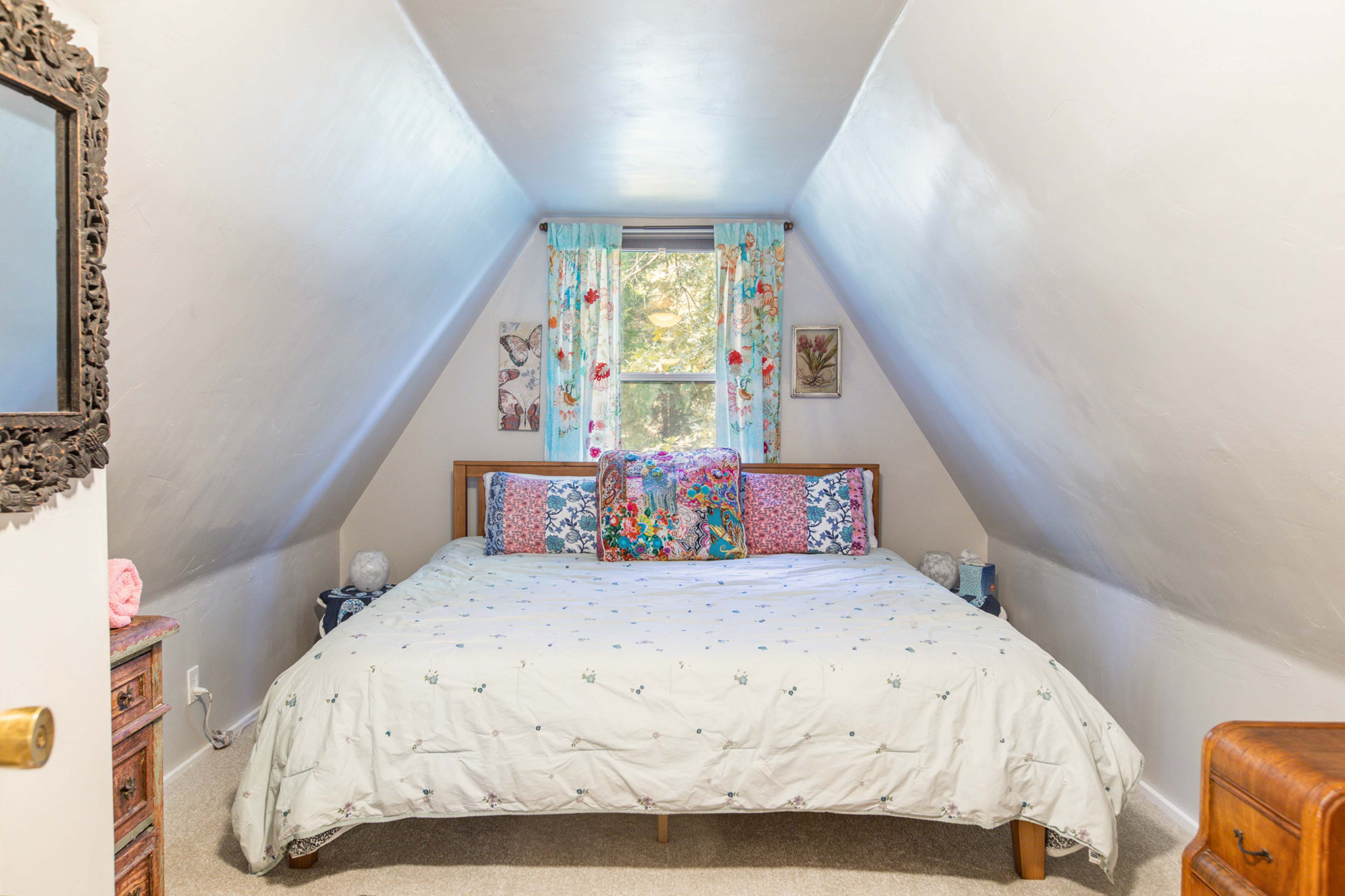The image shows a cozy attic bedroom with a double bed, patterned pillows, and a window adorned with floral curtains.