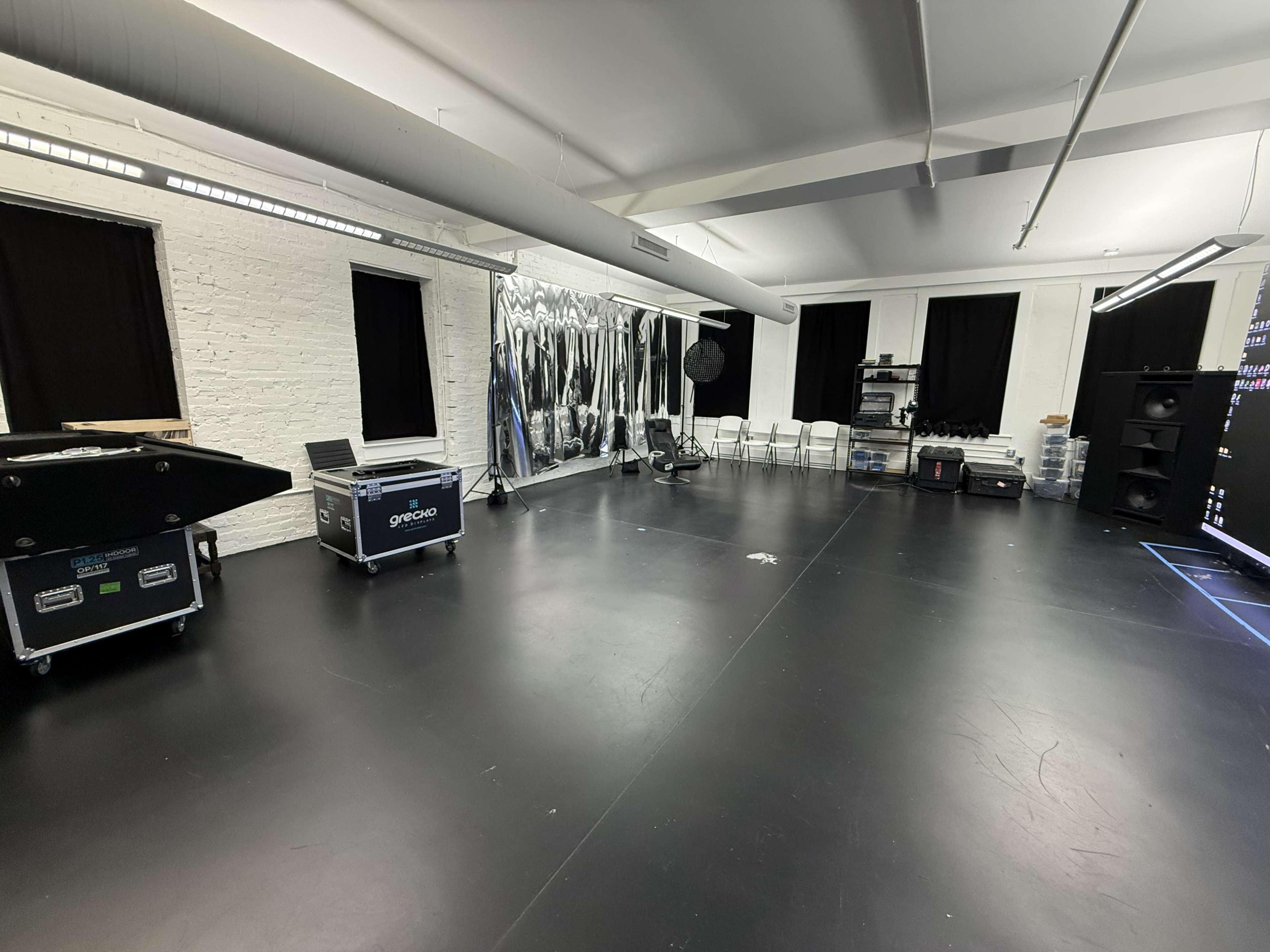 The image shows a spacious studio with black flooring, white brick walls, and several empty chairs arranged in the corner alongside various equipment cases and a mirrored wall.