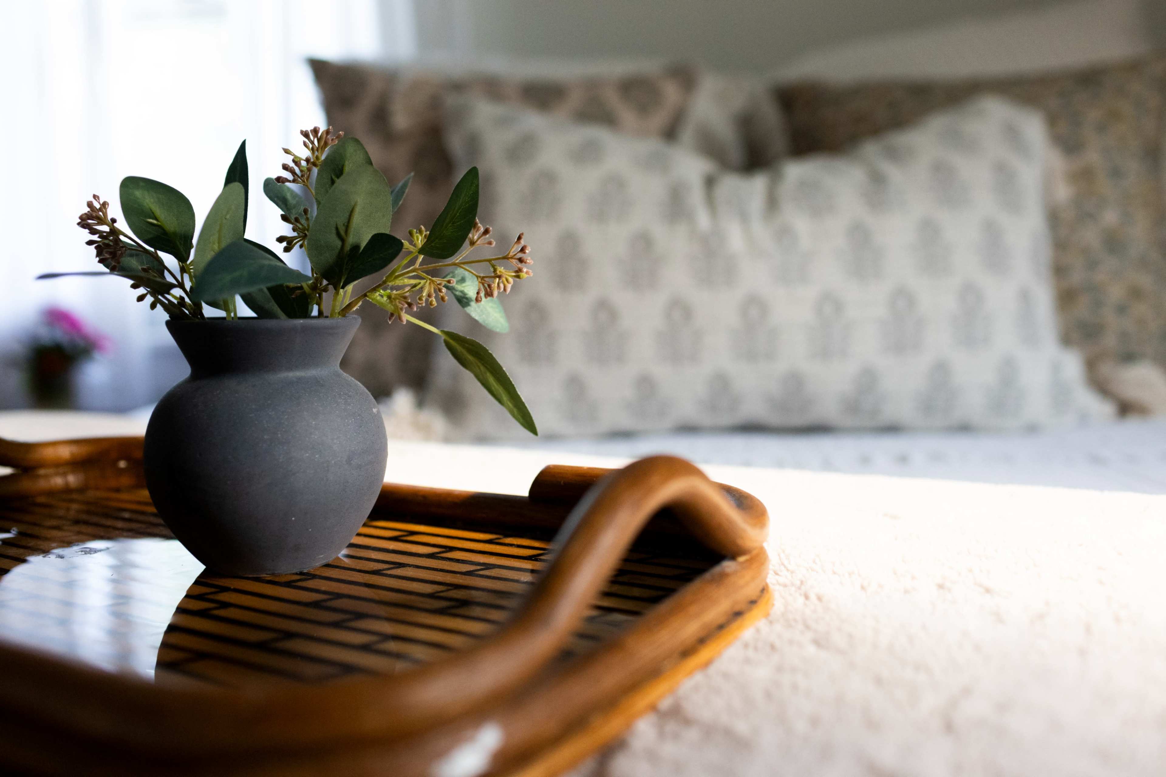 A small black vase with greenery is placed on a wooden tray in a softly lit room with a blurred background featuring a bed and decorative pillows.