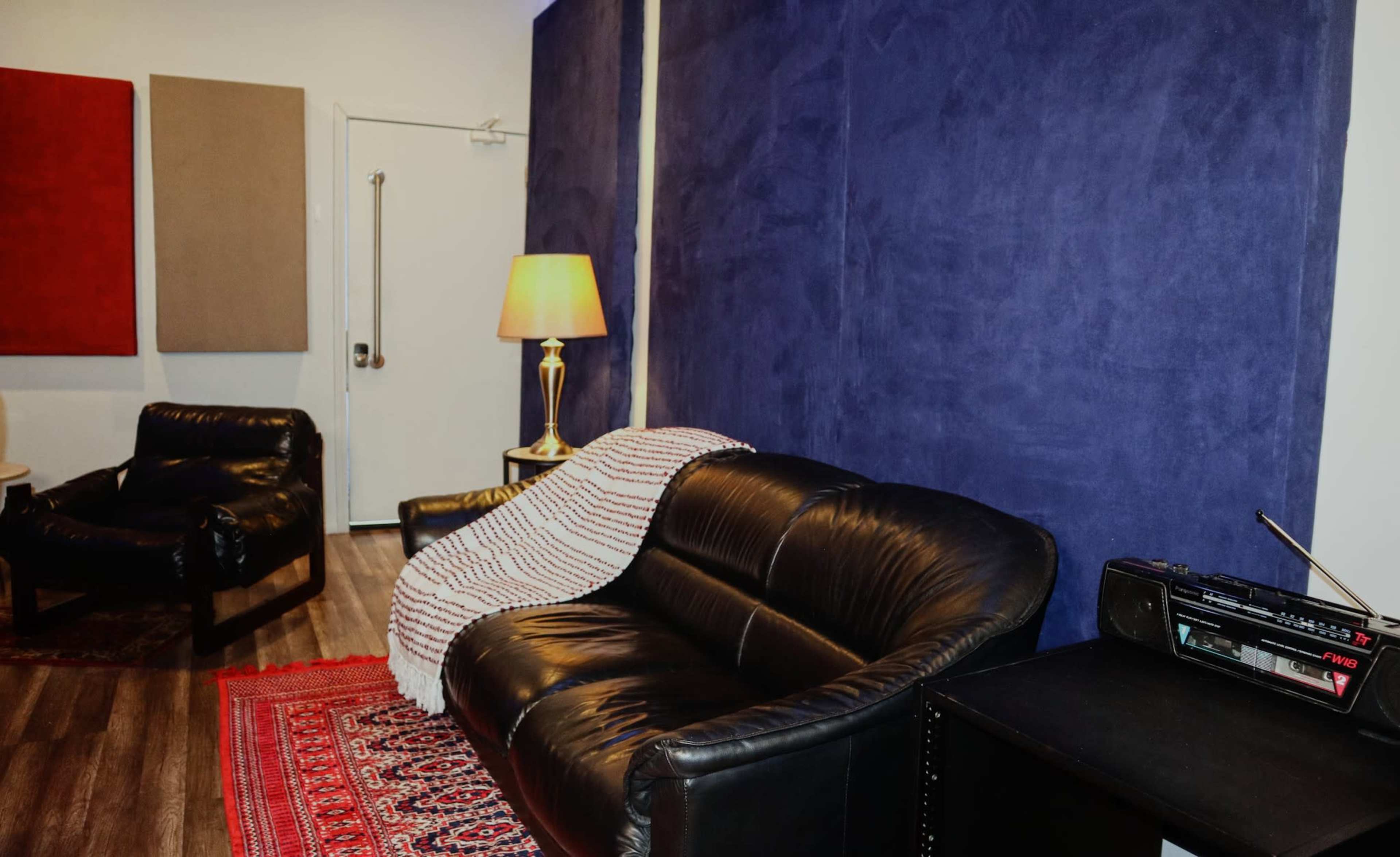 The image shows a cozy room featuring a black leather couch with a draped blanket, a lamp, and colorful wall panels.