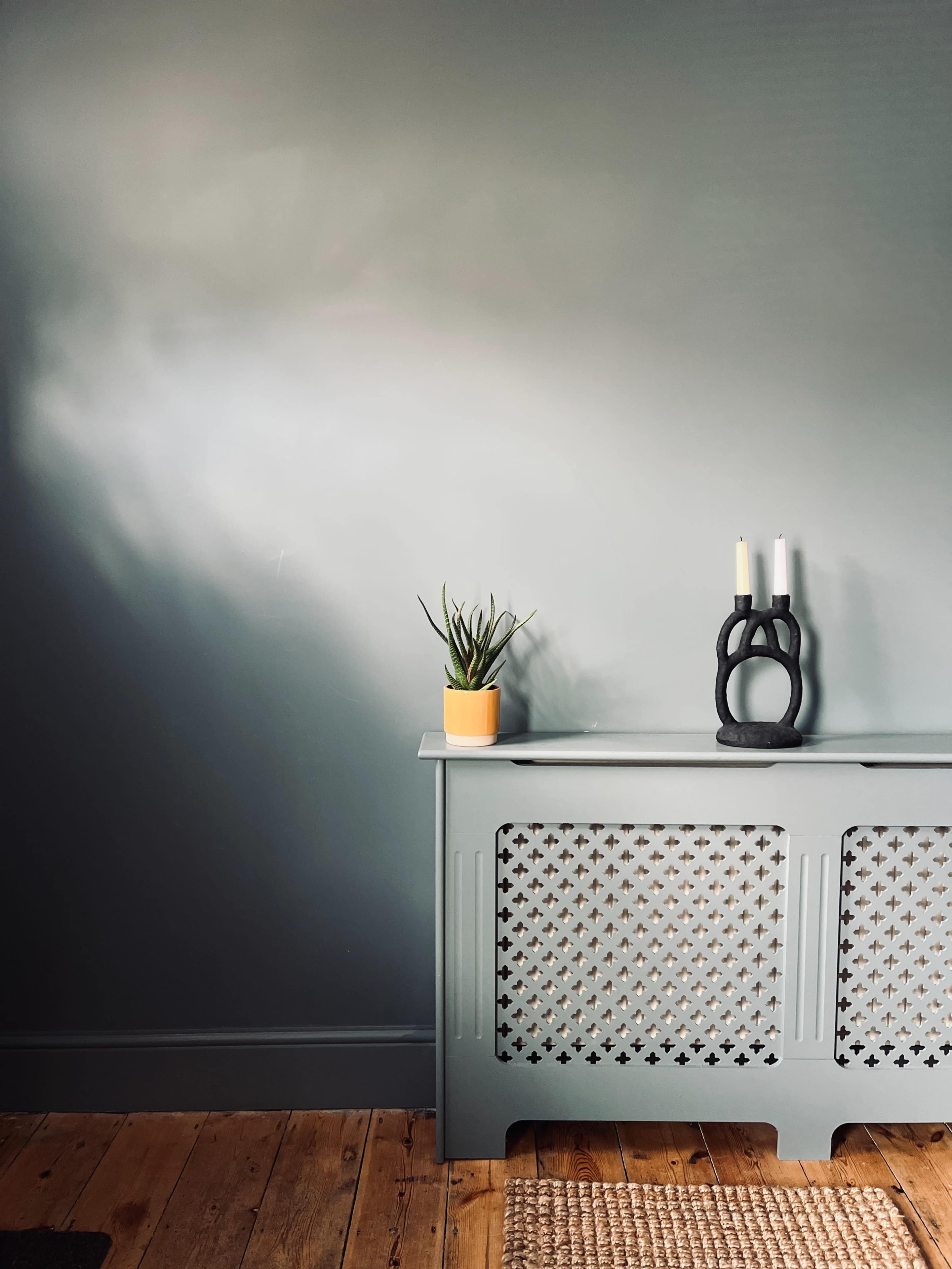 A plant sits in a pot on a console table next to a decorative sculpture and two candles, against a gray wall.