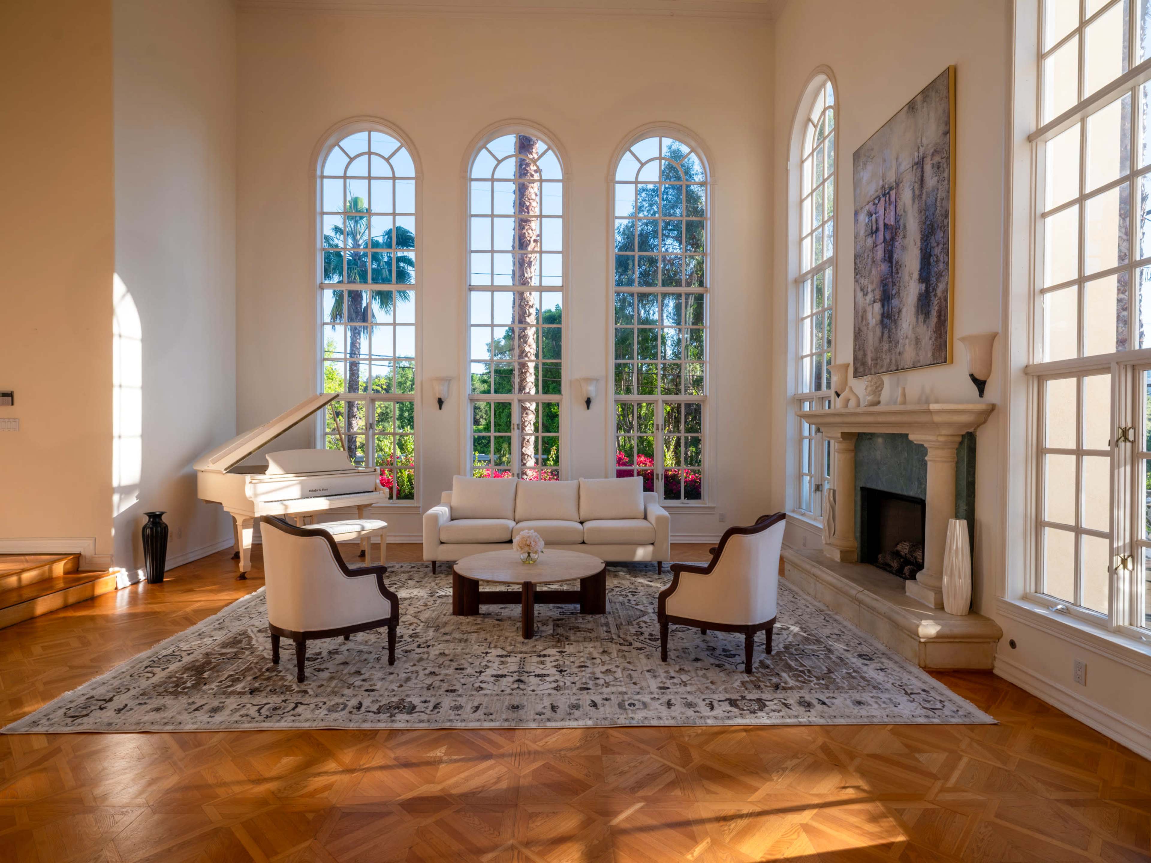 The image shows a bright, spacious living room featuring large arched windows, a white sofa, two chairs, a circular coffee table, and a grand piano, with a view of palm trees and lush greenery outside.