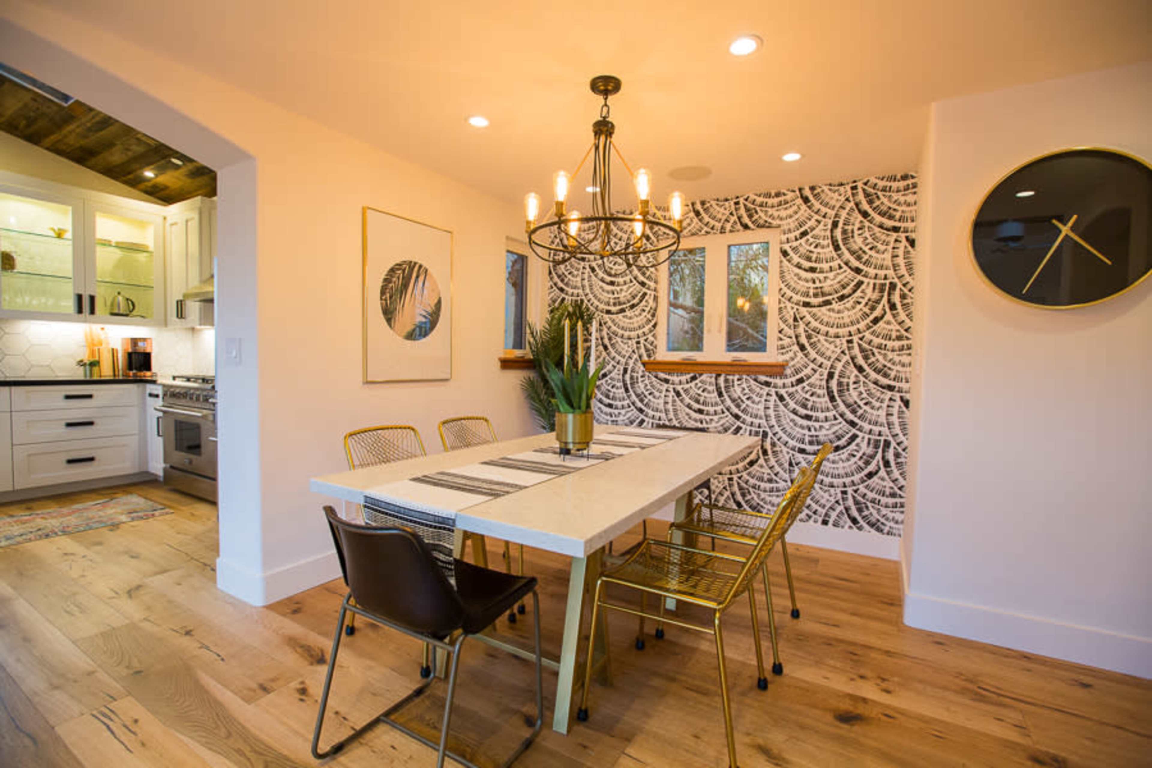 A dining area features a rectangular table surrounded by chairs, with a patterned wall, a chandelier overhead, and a view of the kitchen in the background.