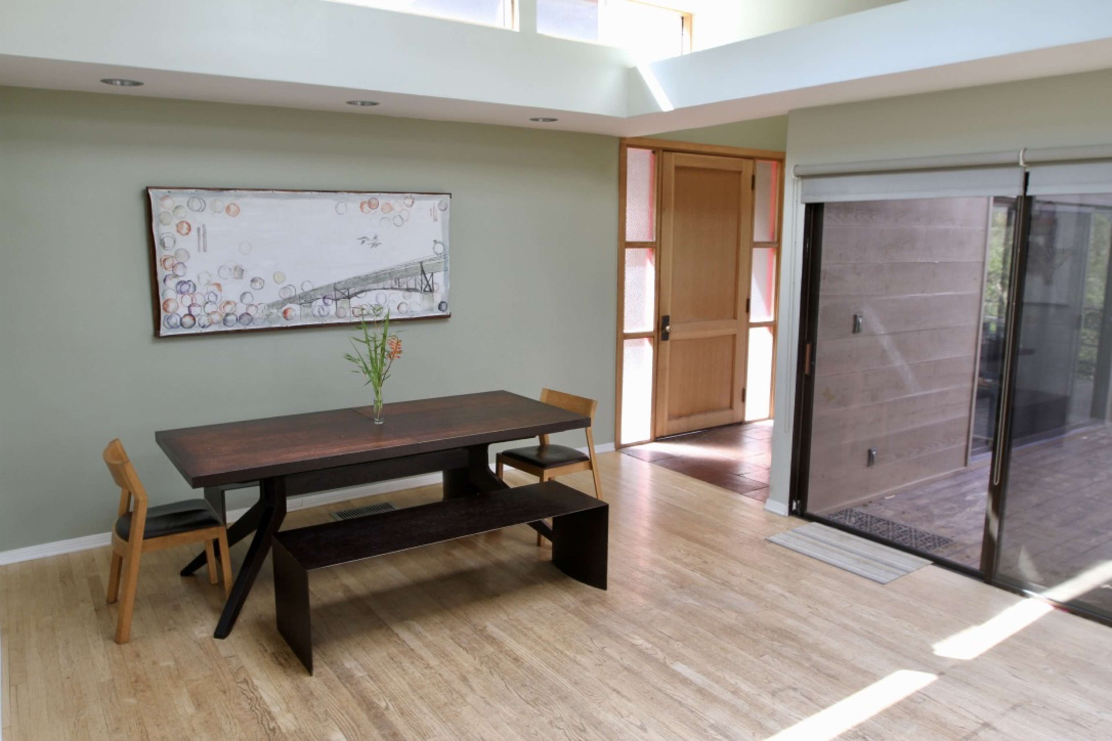 A modern dining area with a wooden table and benches, a framed artwork on the wall, and large glass doors leading to an outdoor space.