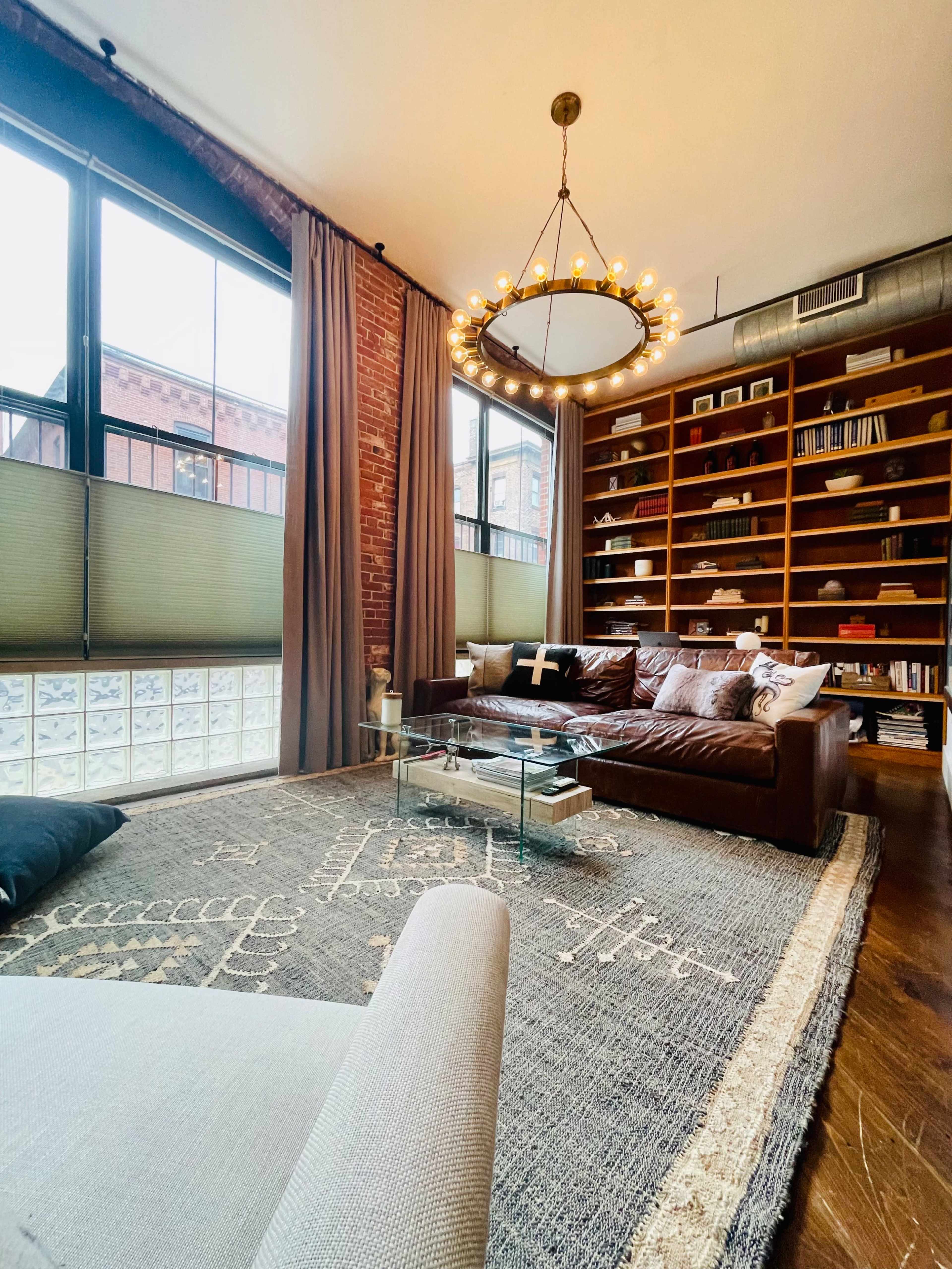 A modern living room featuring a brown leather sofa, a circular chandelier, large windows with light-filtering shades, a glass coffee table, and a bookshelf filled with books.