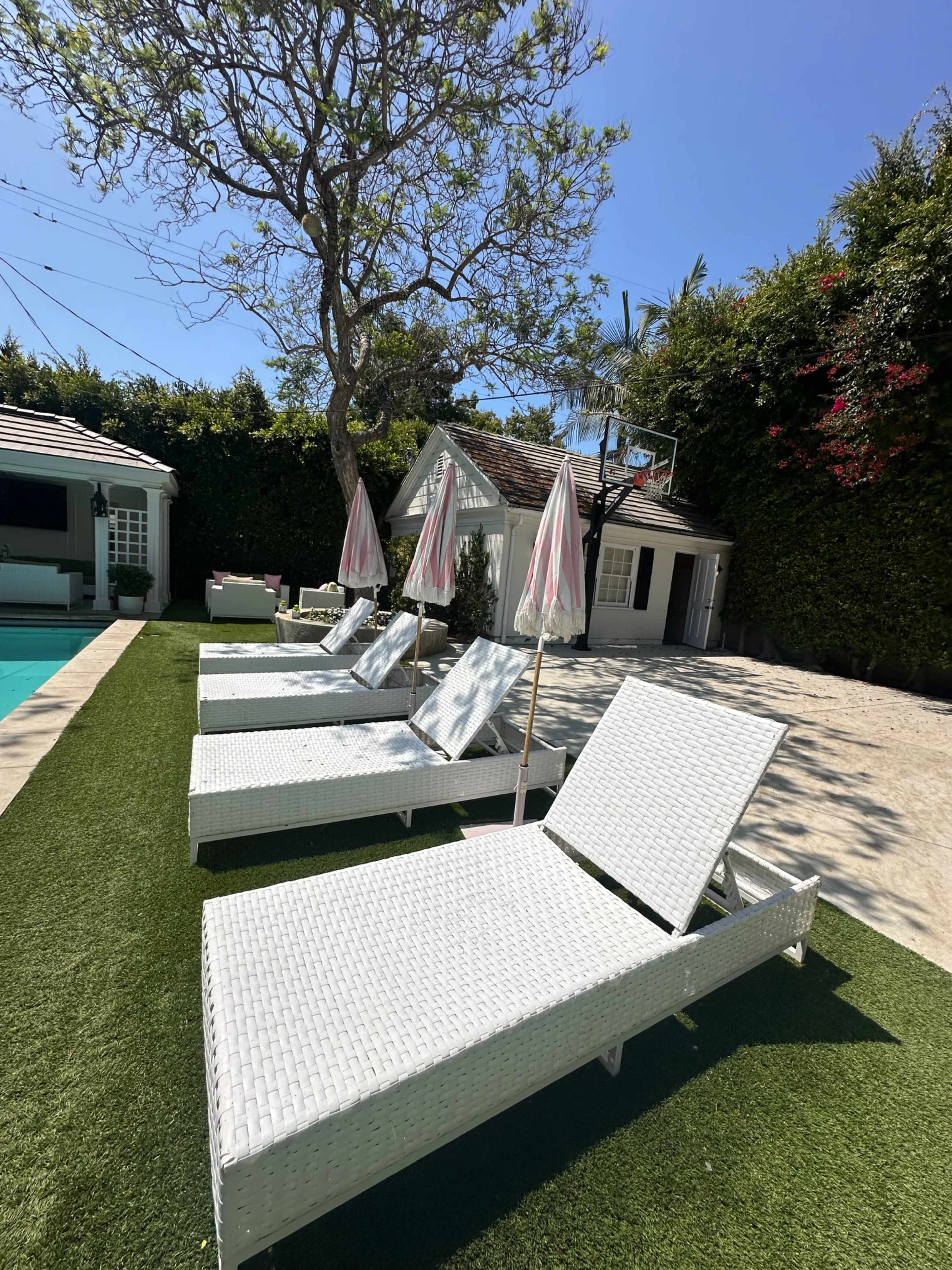 The image shows a poolside area with three white lounge chairs and pink umbrellas set on green grass next to a house surrounded by trees and shrubs.