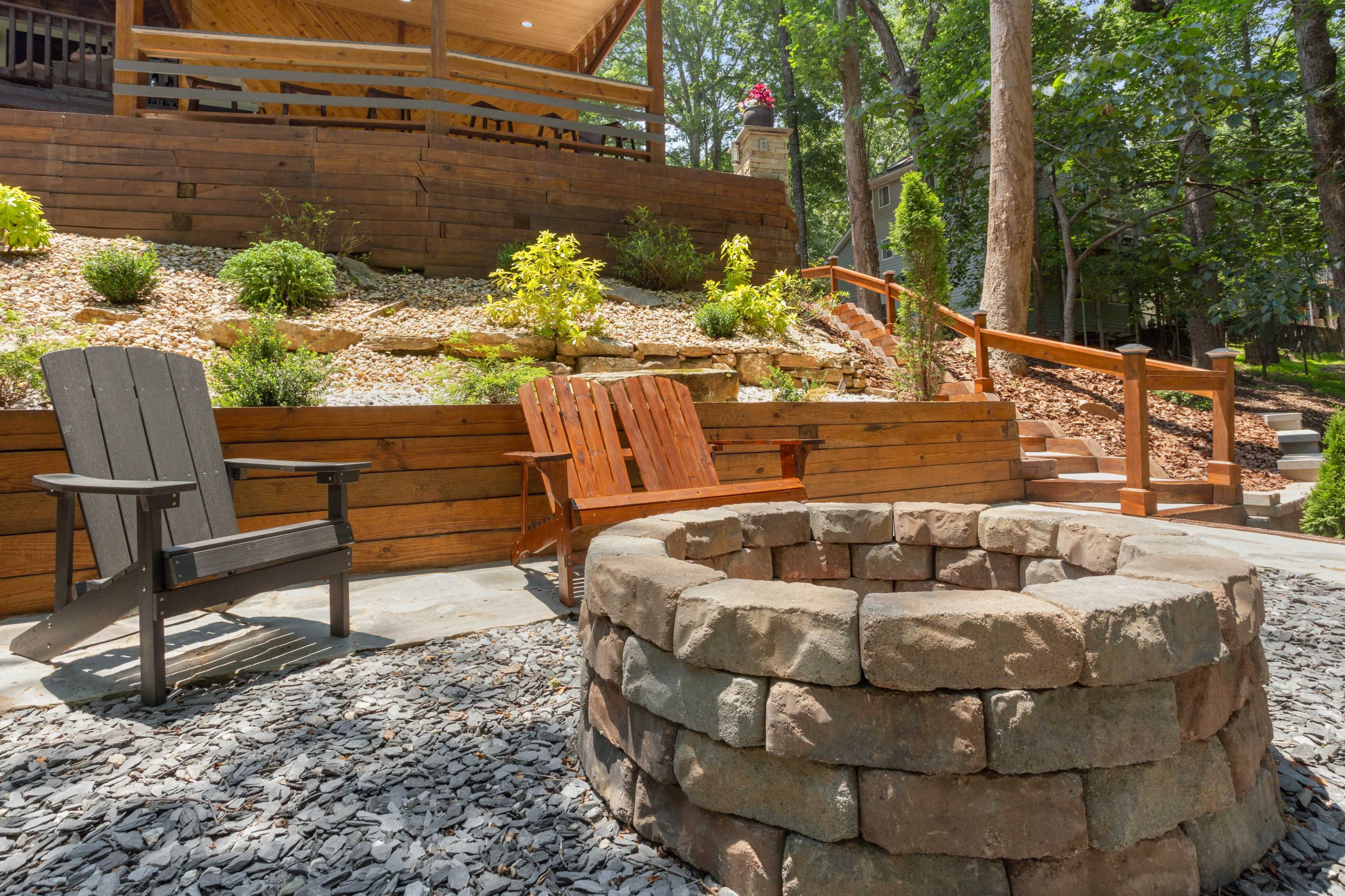 The image shows a backyard setting featuring a stone fire pit surrounded by gravel, with wooden seating and a landscaped area in the background.
