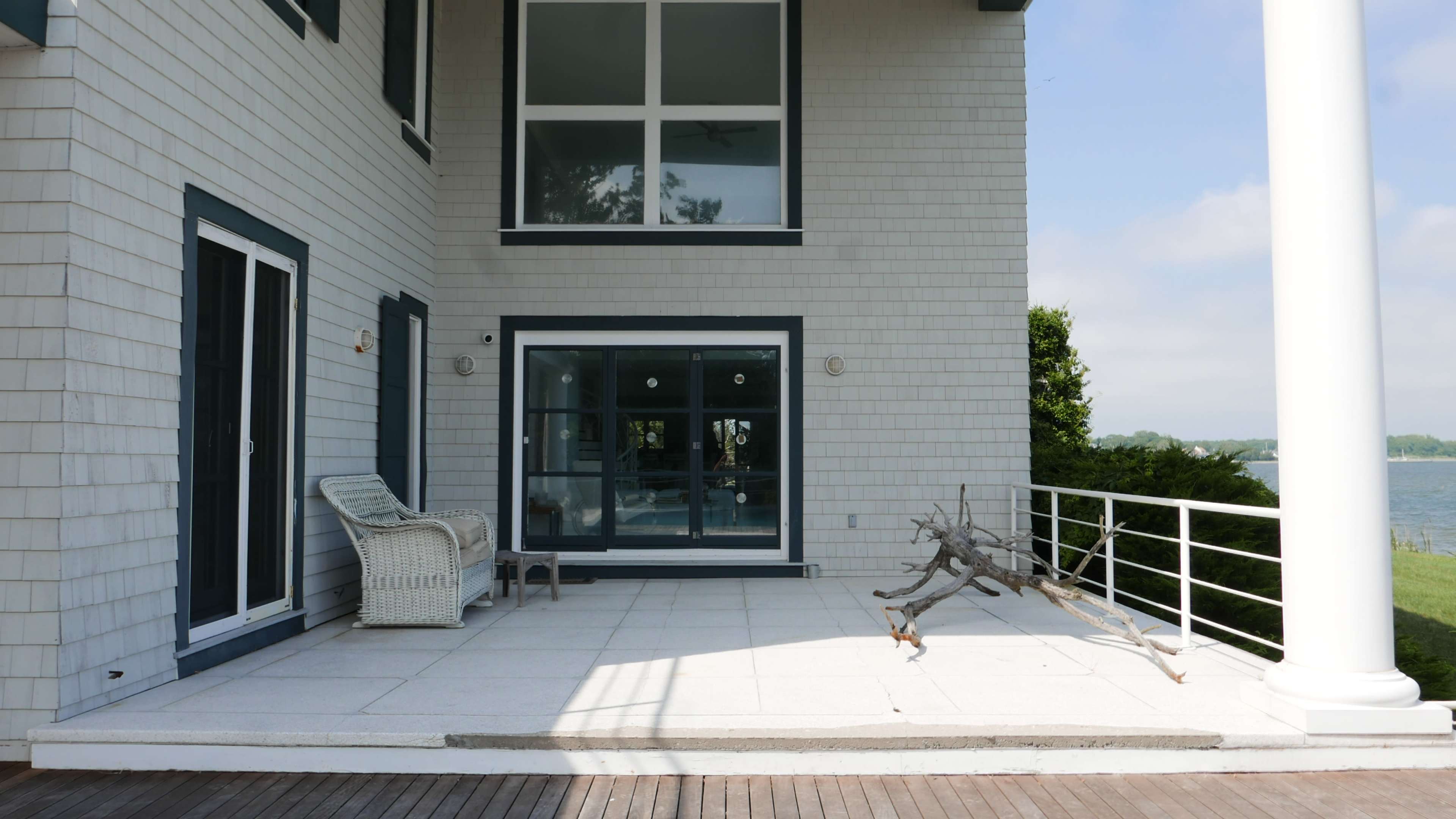 A modern patio area with a wicker chair, large windows, and a section of wood decking overlooking a body of water.