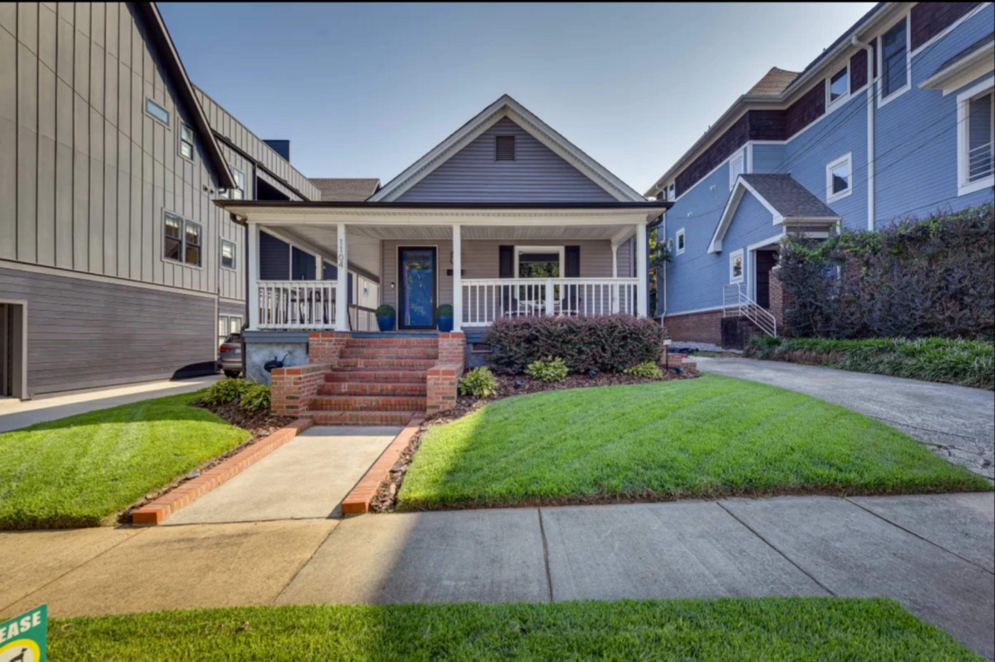 A single-story house with a front porch and brick steps, situated on a grassy lawn between two larger buildings.