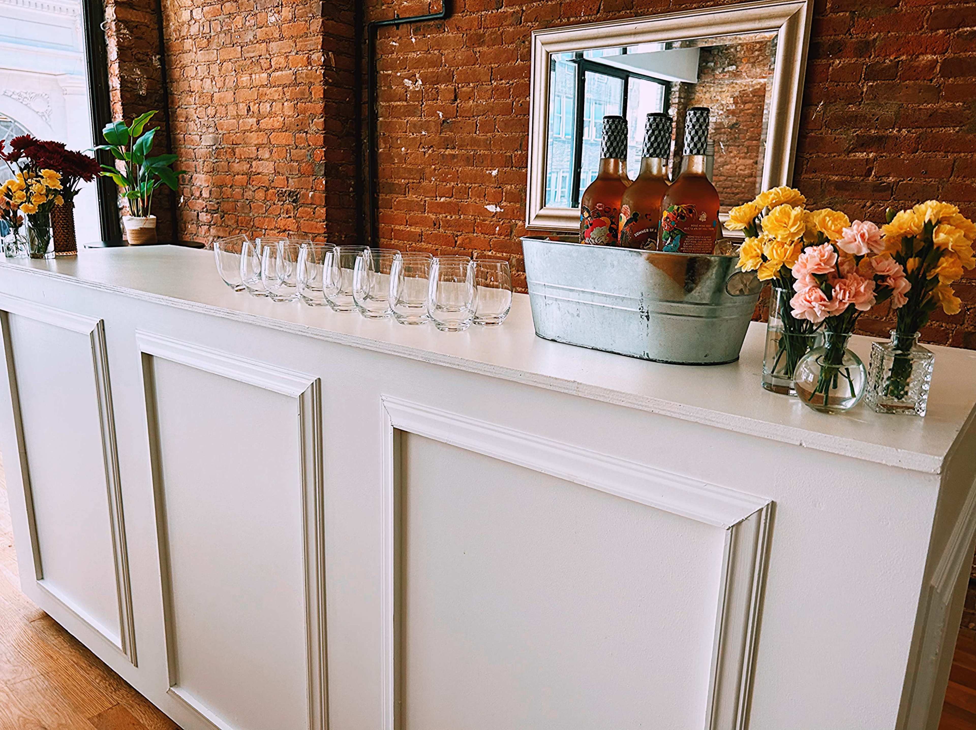 A white bar with a mirrored back features a metal bucket holding several bottles, surrounded by glasses and flower arrangements against a brick wall.