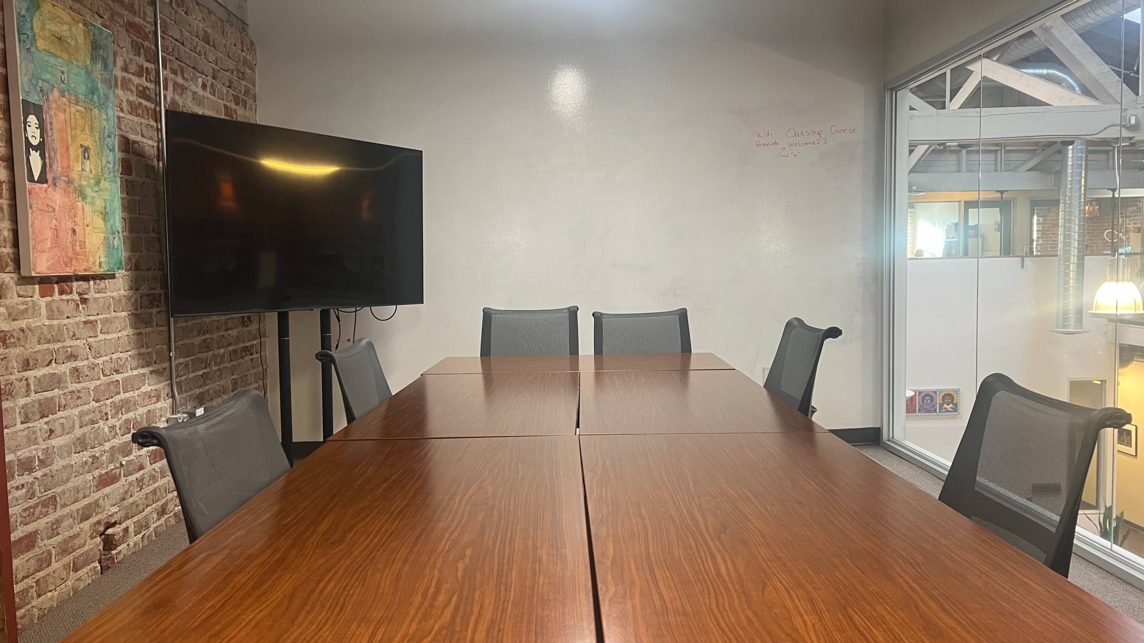 A conference room with a large wooden table surrounded by six chairs, a television mounted on the wall, and a window showing an indoor space.