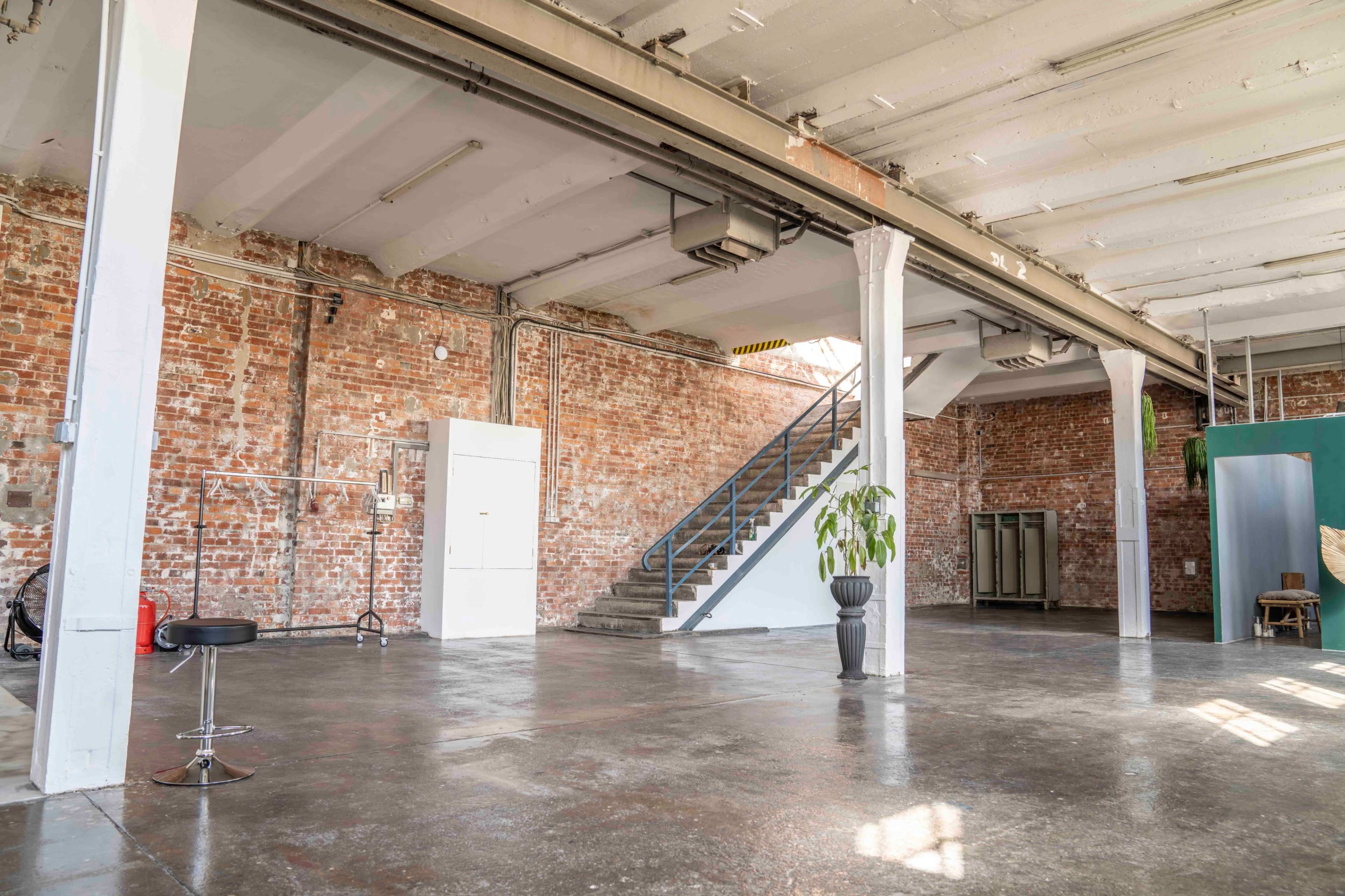 The image shows a spacious industrial-style room with exposed brick walls, concrete floors, and a staircase leading to an upper level.
