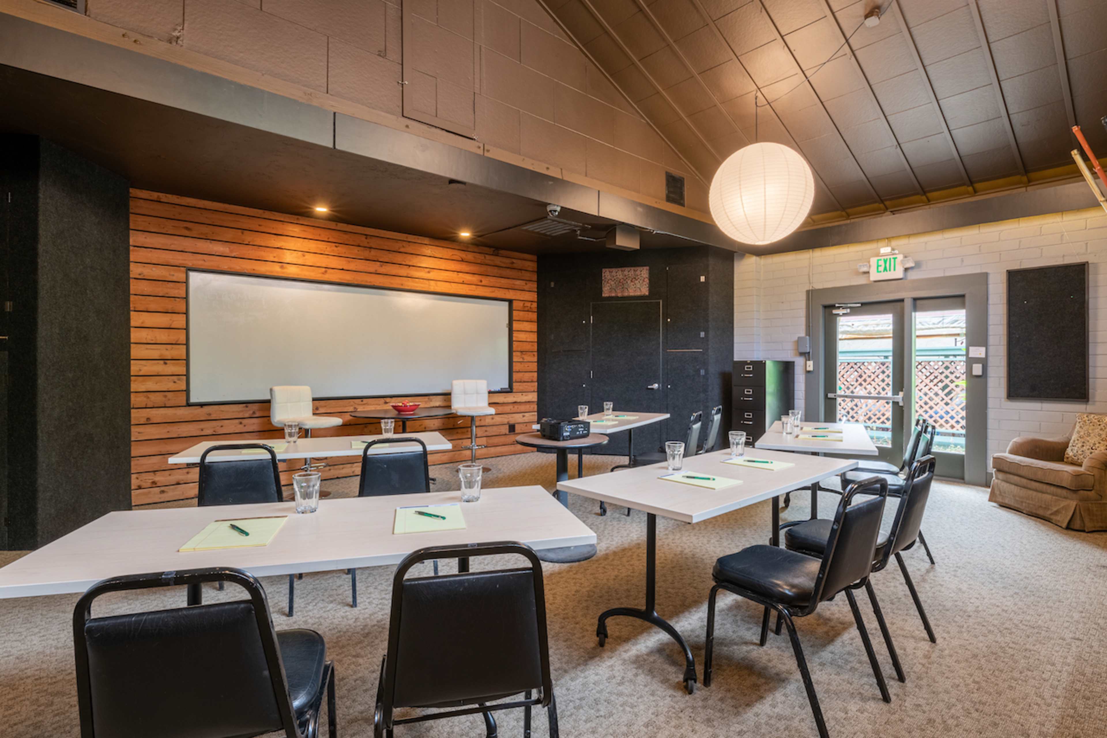 A meeting room with several tables and chairs arranged for a presentation, featuring a whiteboard and a large pendant light overhead.
