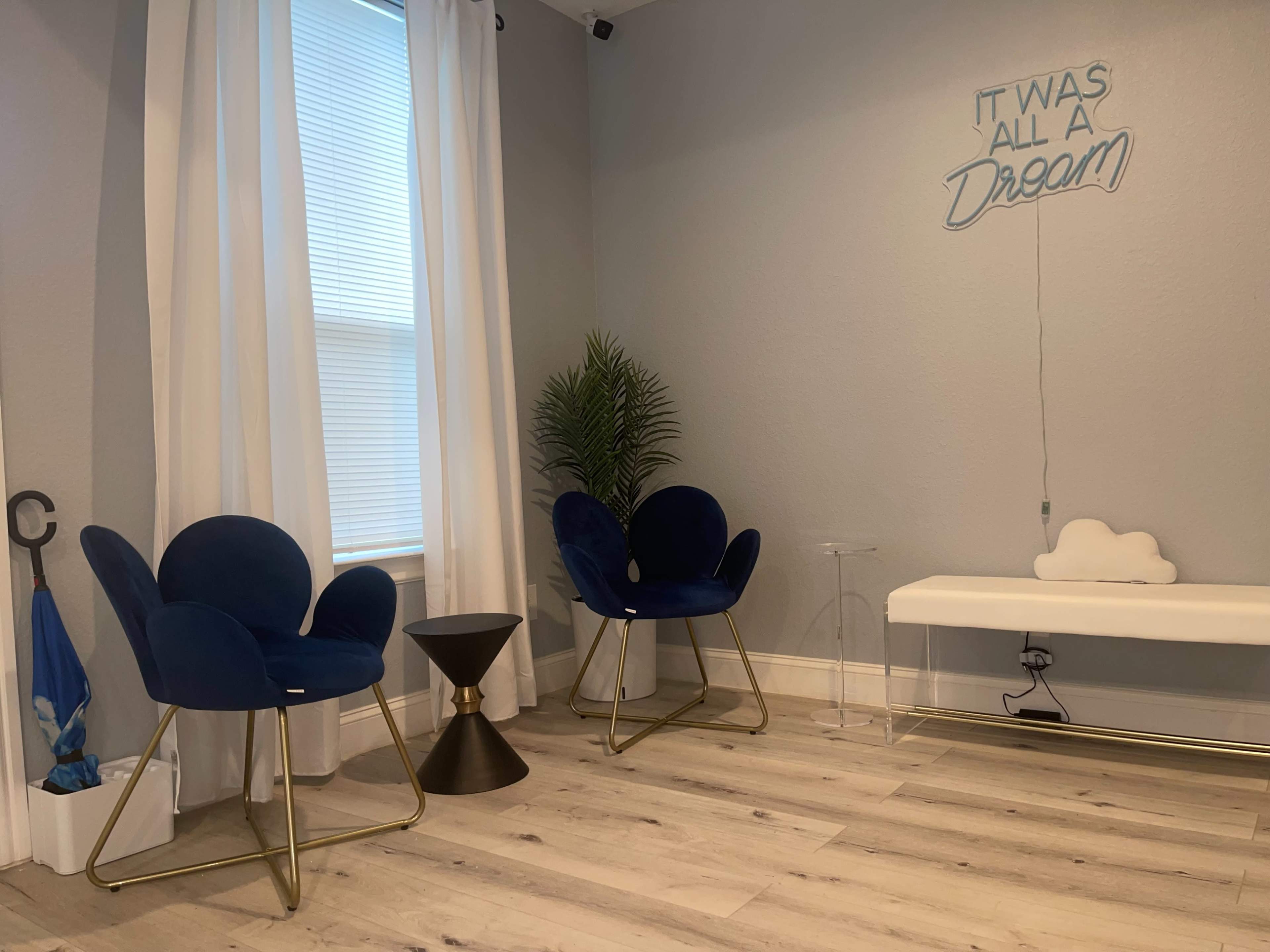 A minimalist room featuring two blue chairs, a small black side table, and a white bench, with a decorative cloud and a wall sign that reads "IT WAS ALL A DREAM."