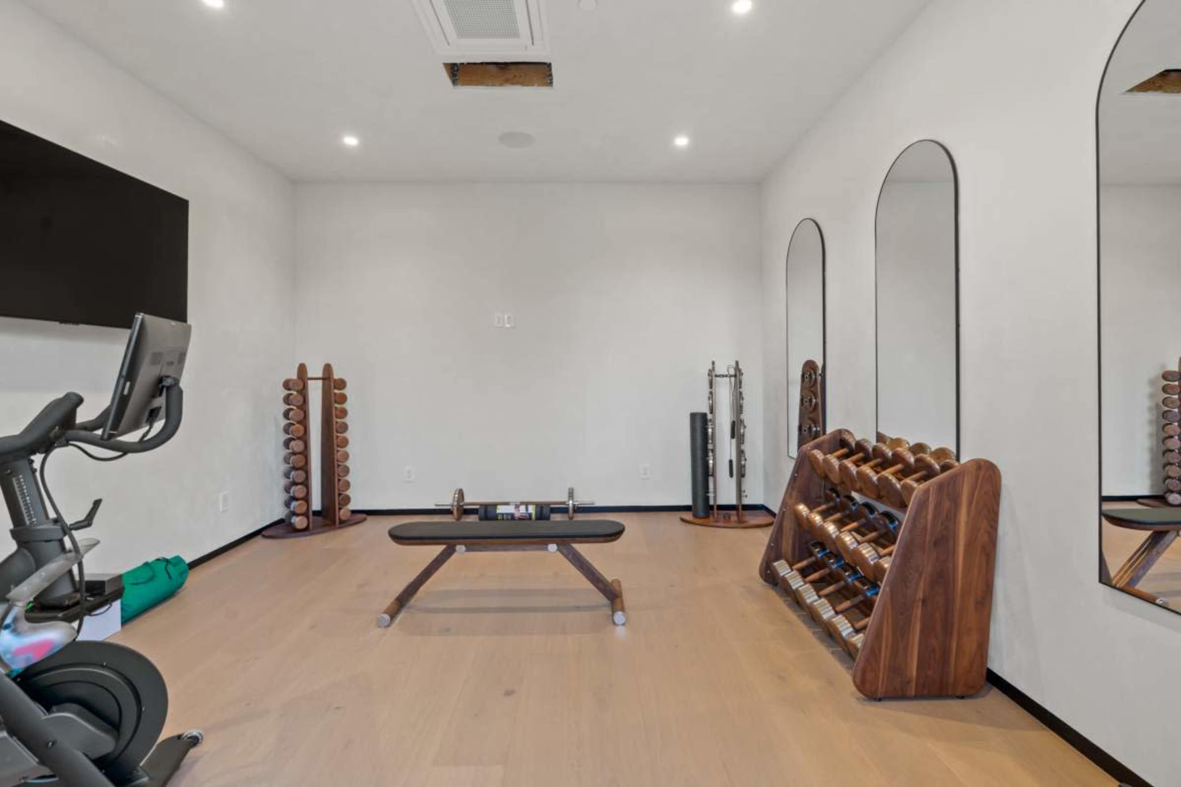 The image shows a modern home gym with a weight bench, dumbbells on racks, a stationary bike, and mirrors on the walls.