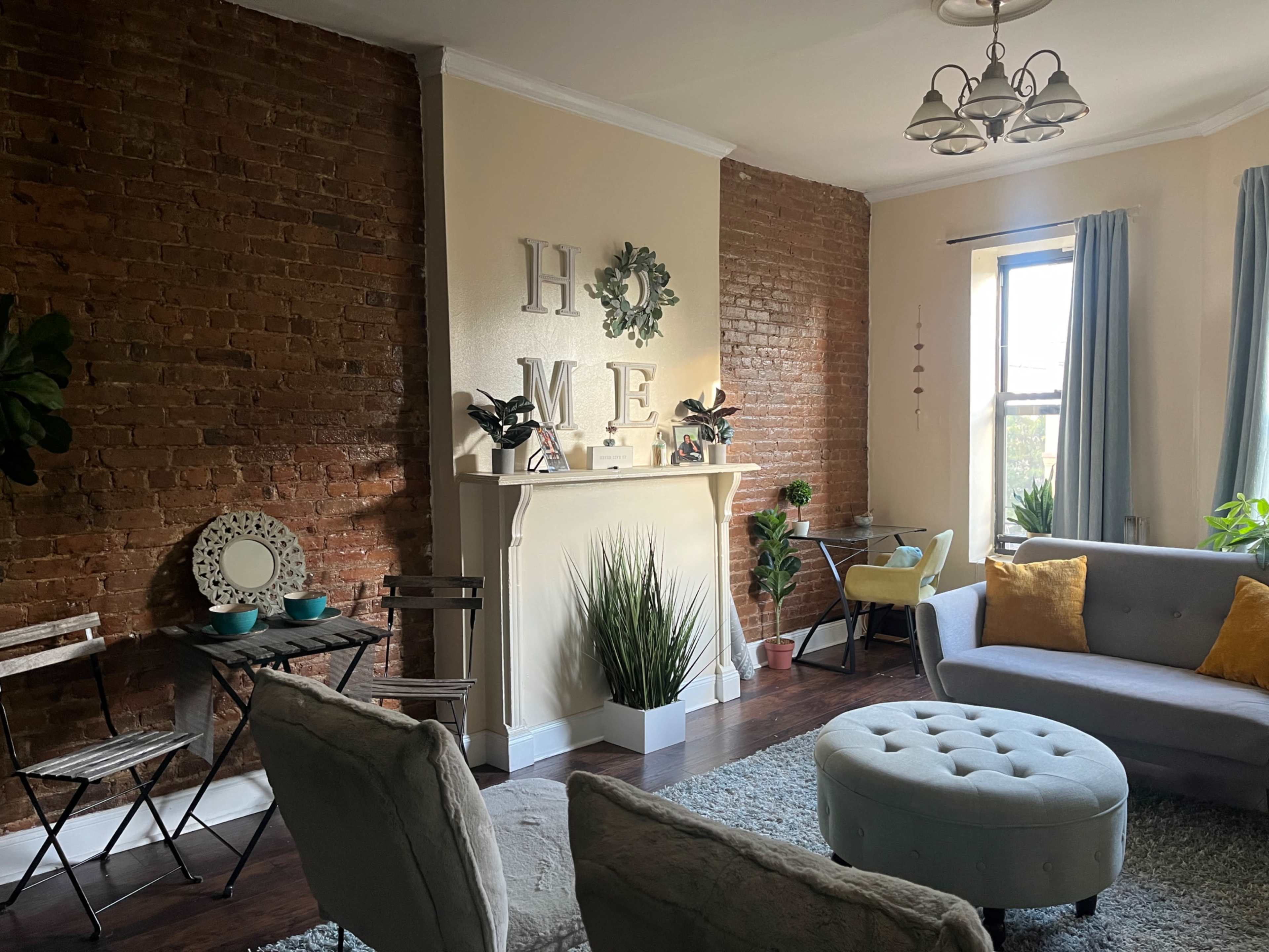 A cozy living room features exposed brick walls, a light-colored mantel decorated with plants, and comfortable seating arranged around a round ottoman.