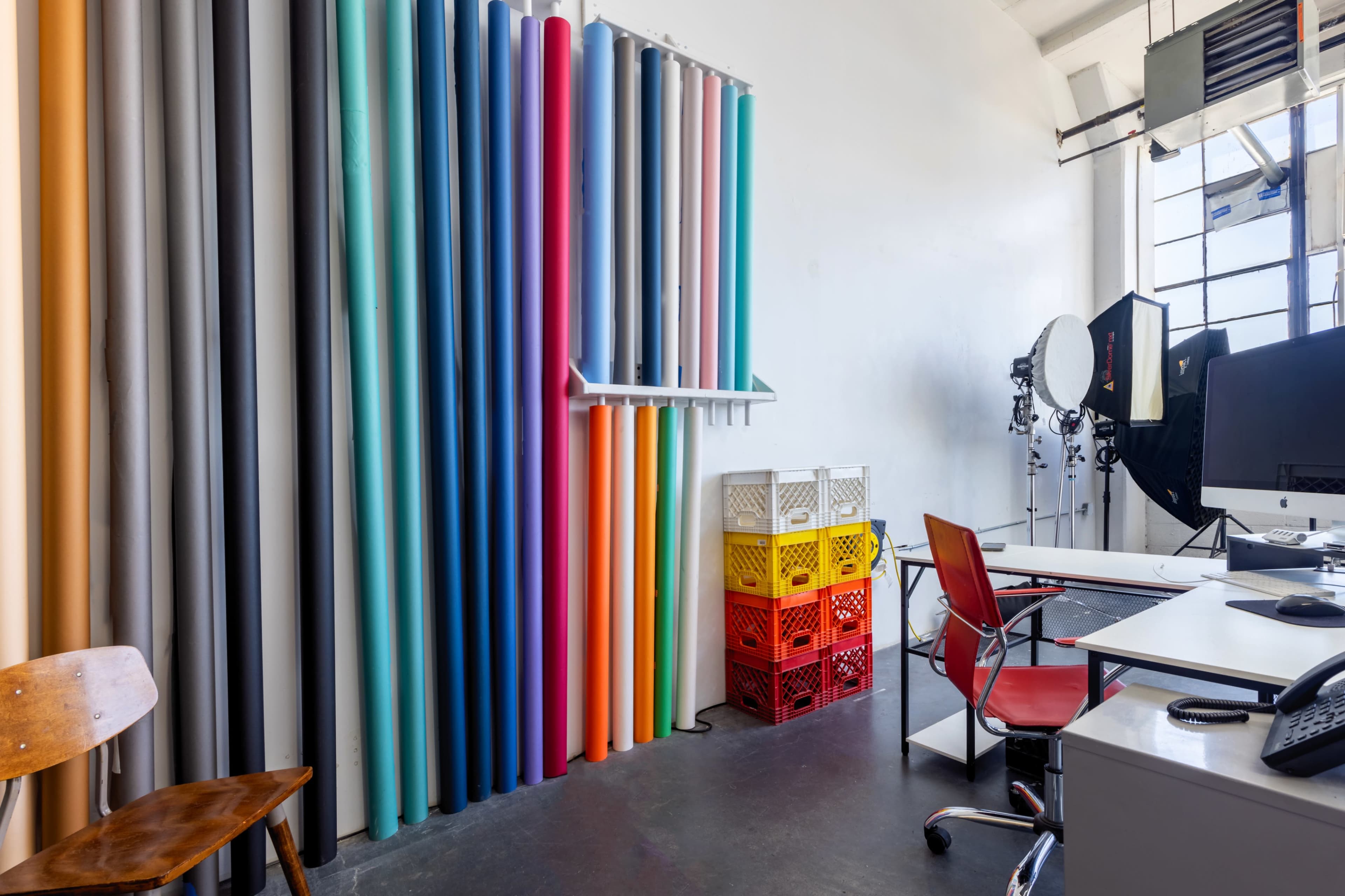 The image shows a photography studio with colorful backdrops, crates, a desk, and a chair.