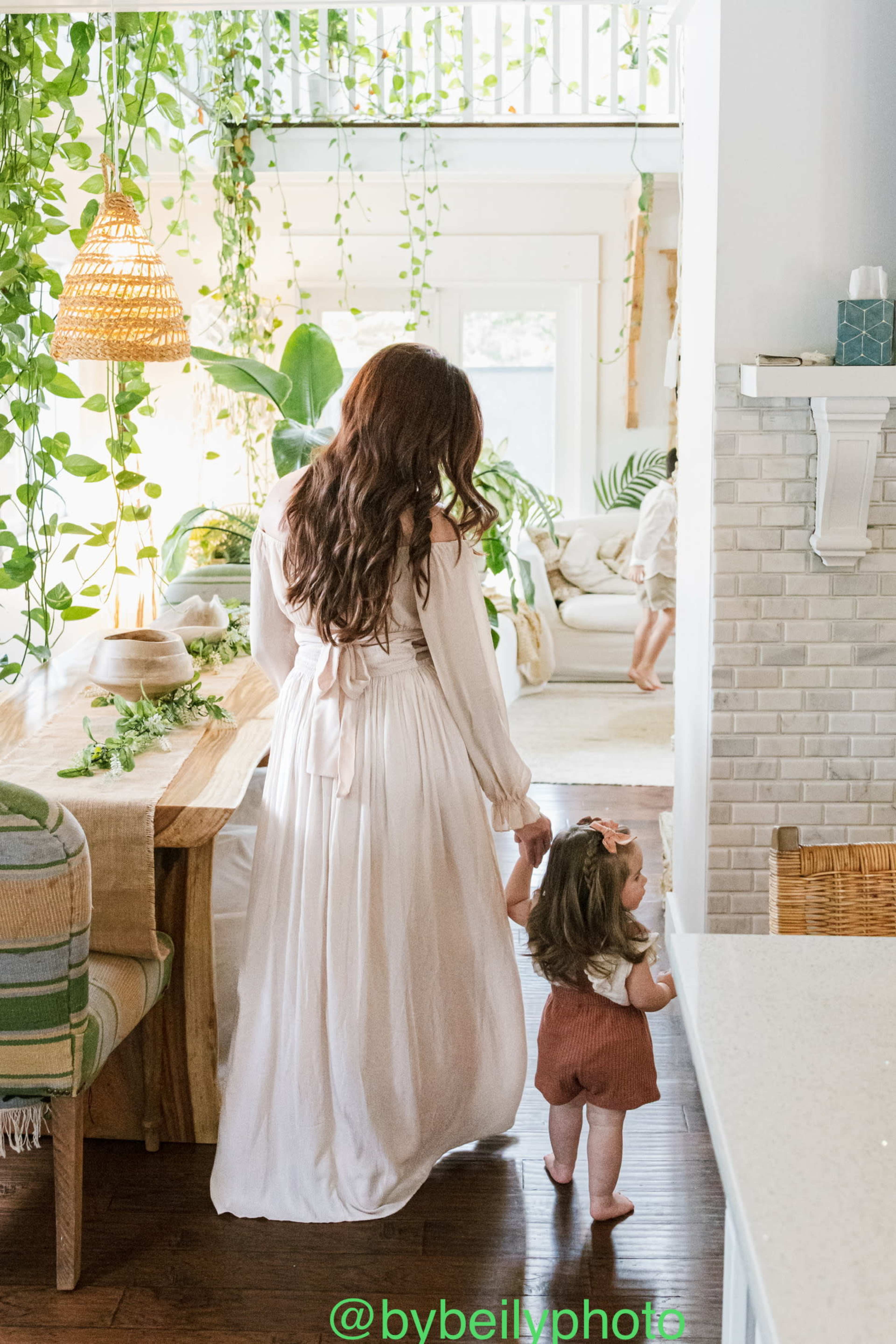 A woman in a flowy dress holds the hand of a small child while walking through a bright, leafy indoor space.