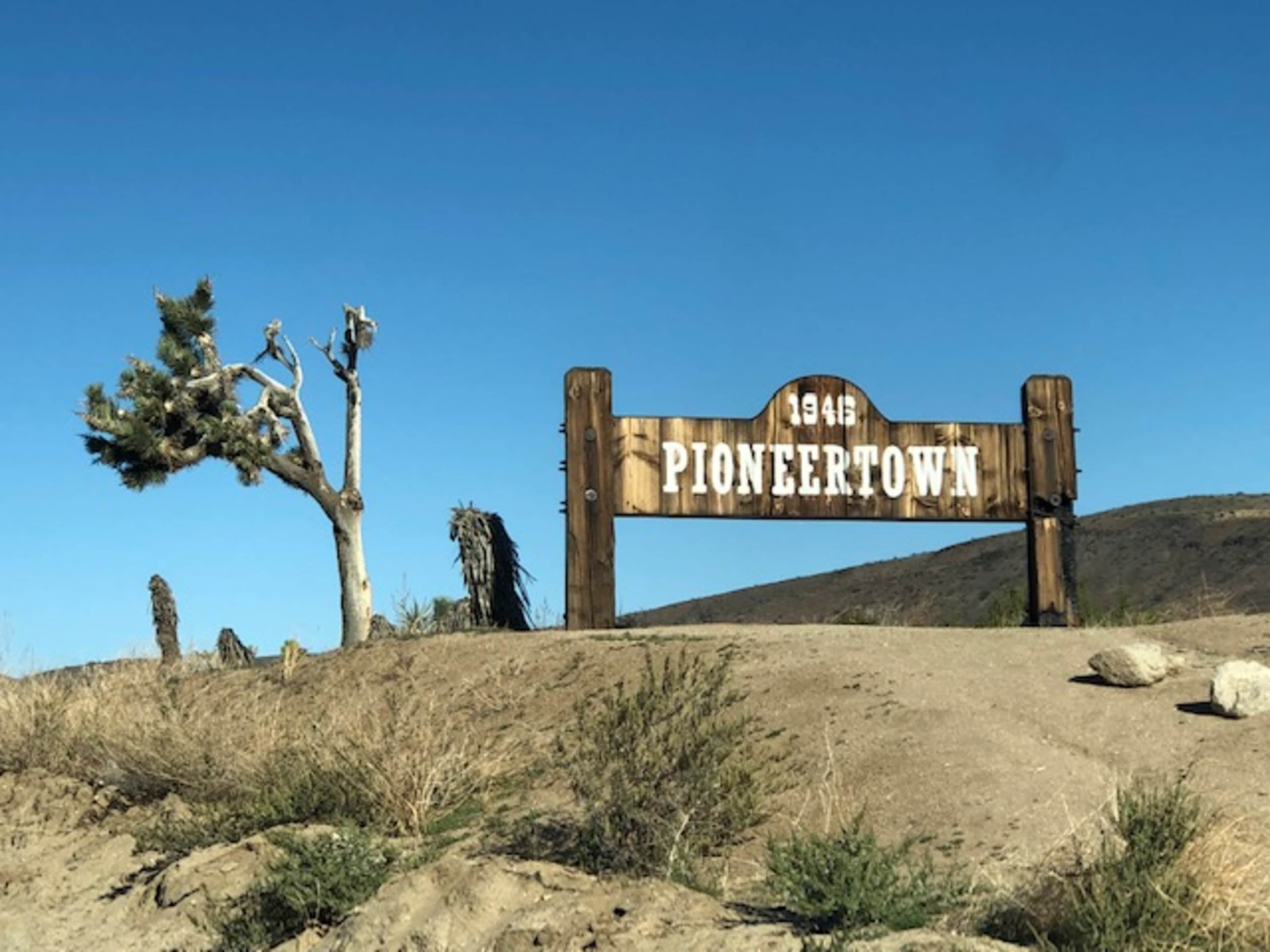 A wooden sign reads "Pioneertown 1946" beside a sparse, dry landscape and a lone tree.