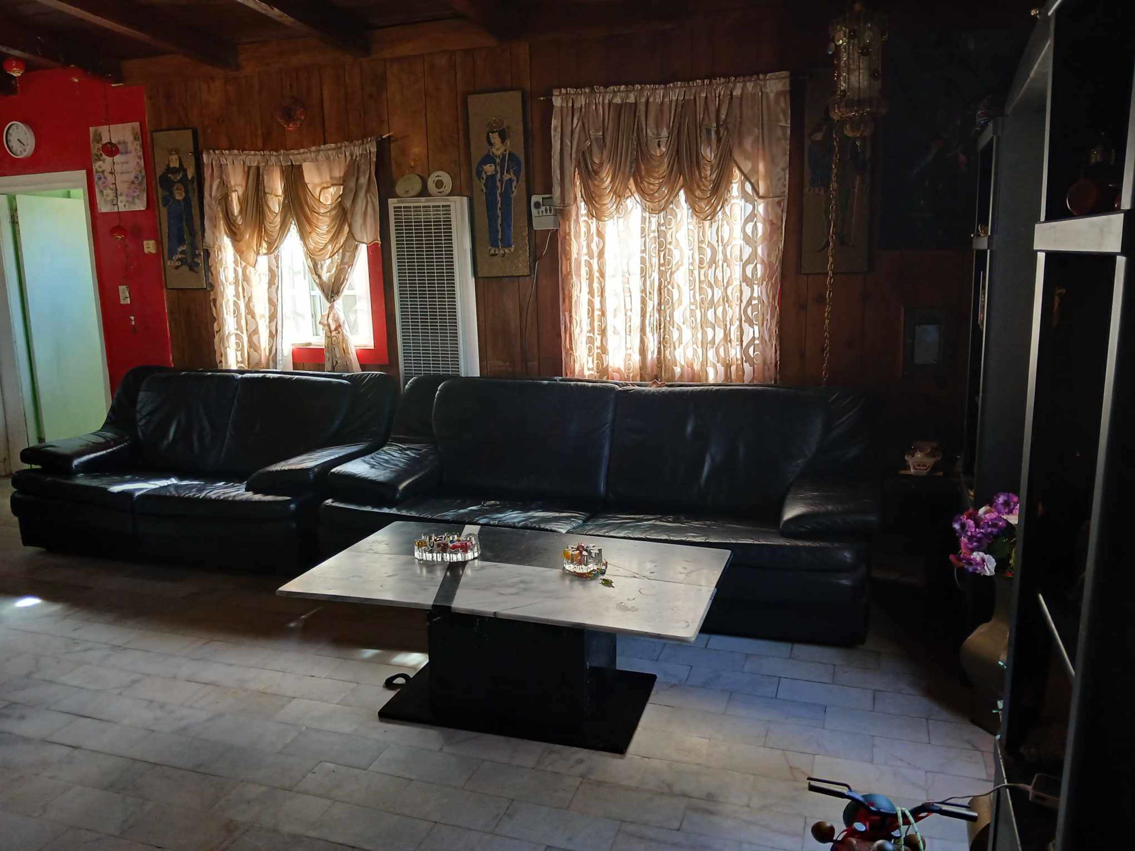 The image shows a living room with two black leather sofas facing a glass coffee table, surrounded by curtains and wooden paneling.
