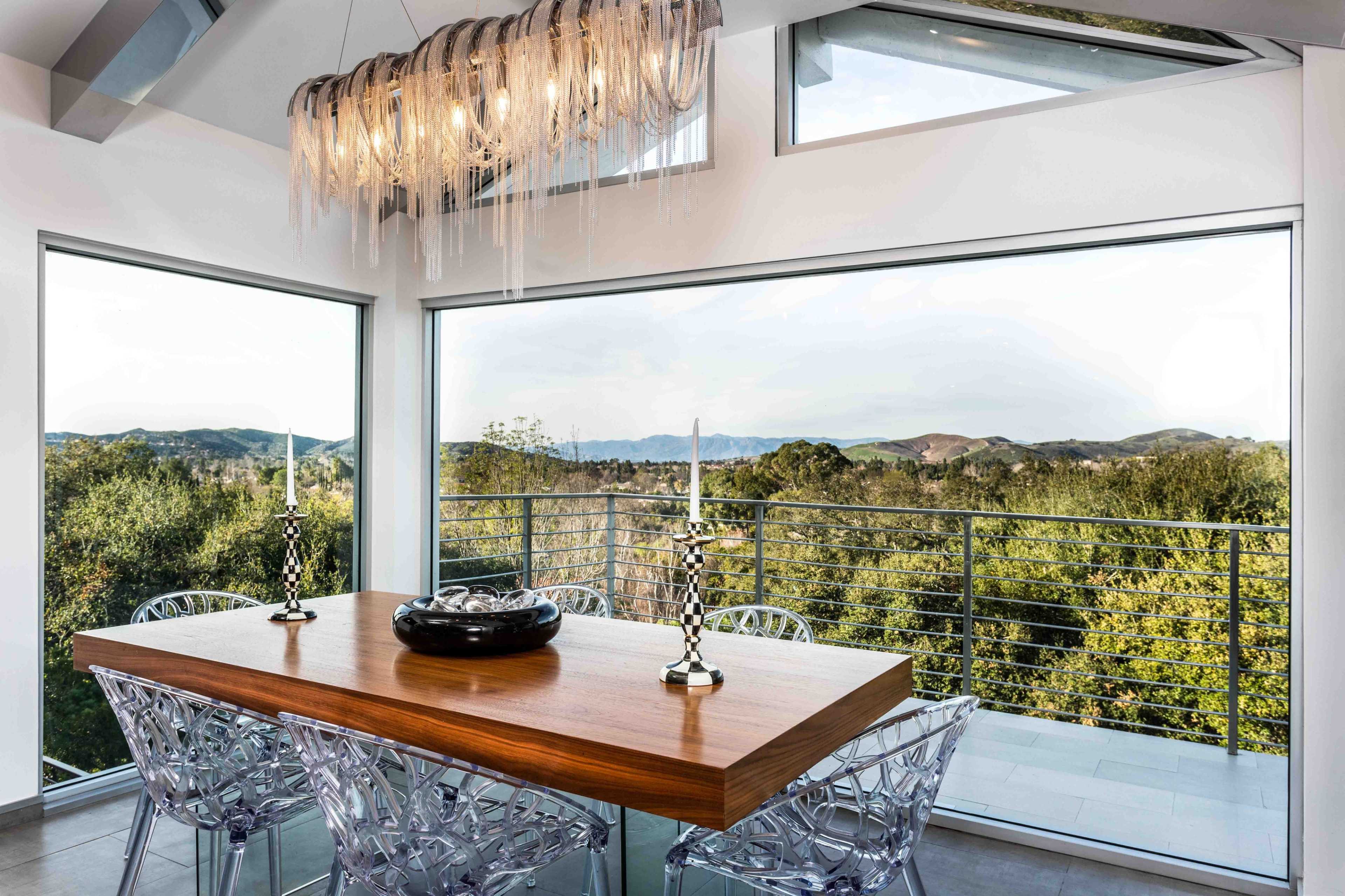 A wooden dining table with clear acrylic chairs is set near large windows that offer a view of hills and trees.