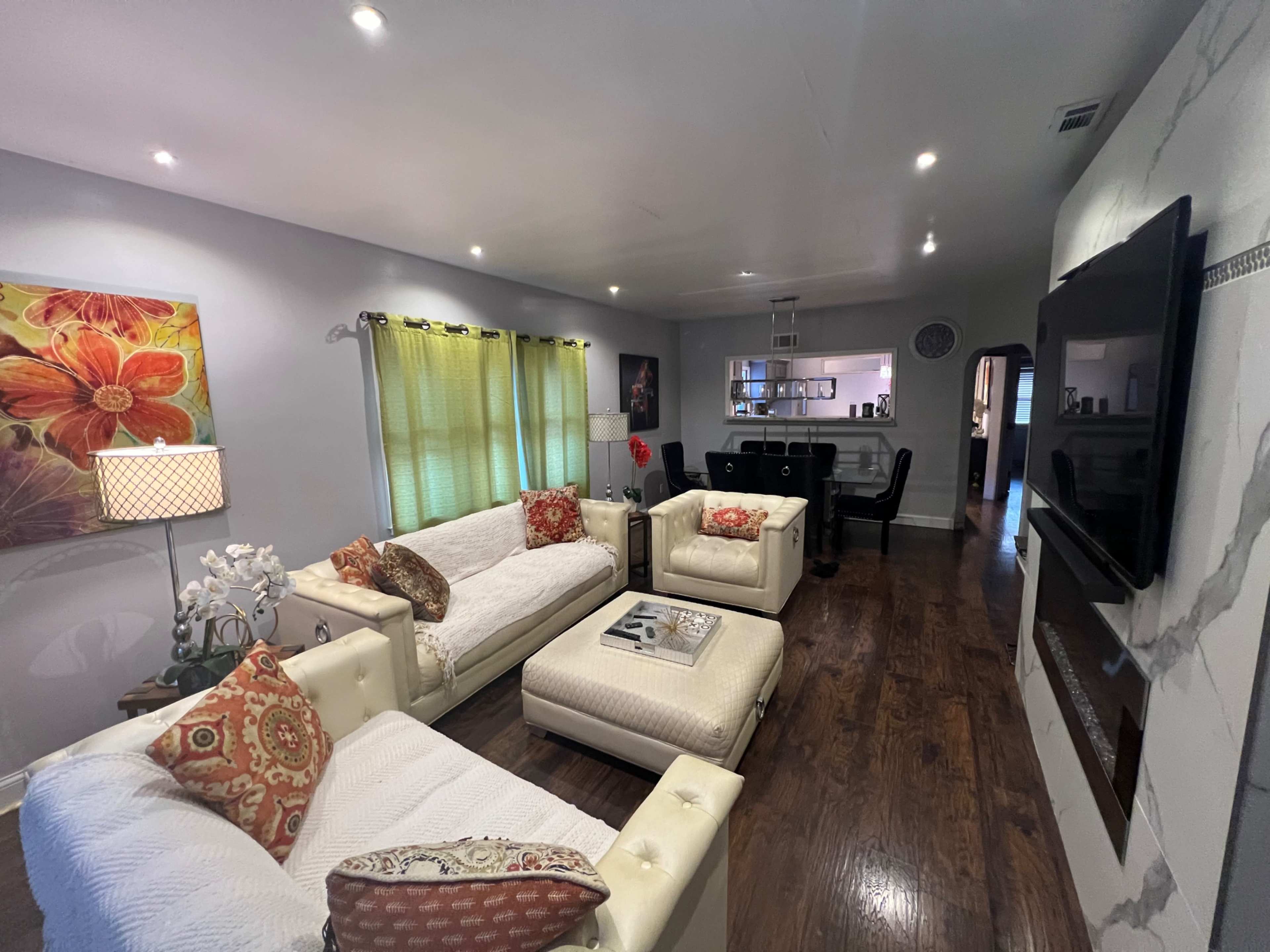 A modern living room features two white sofas, a coffee table, and a large wall-mounted television, with green curtains and decorative artwork adding to the ambiance.
