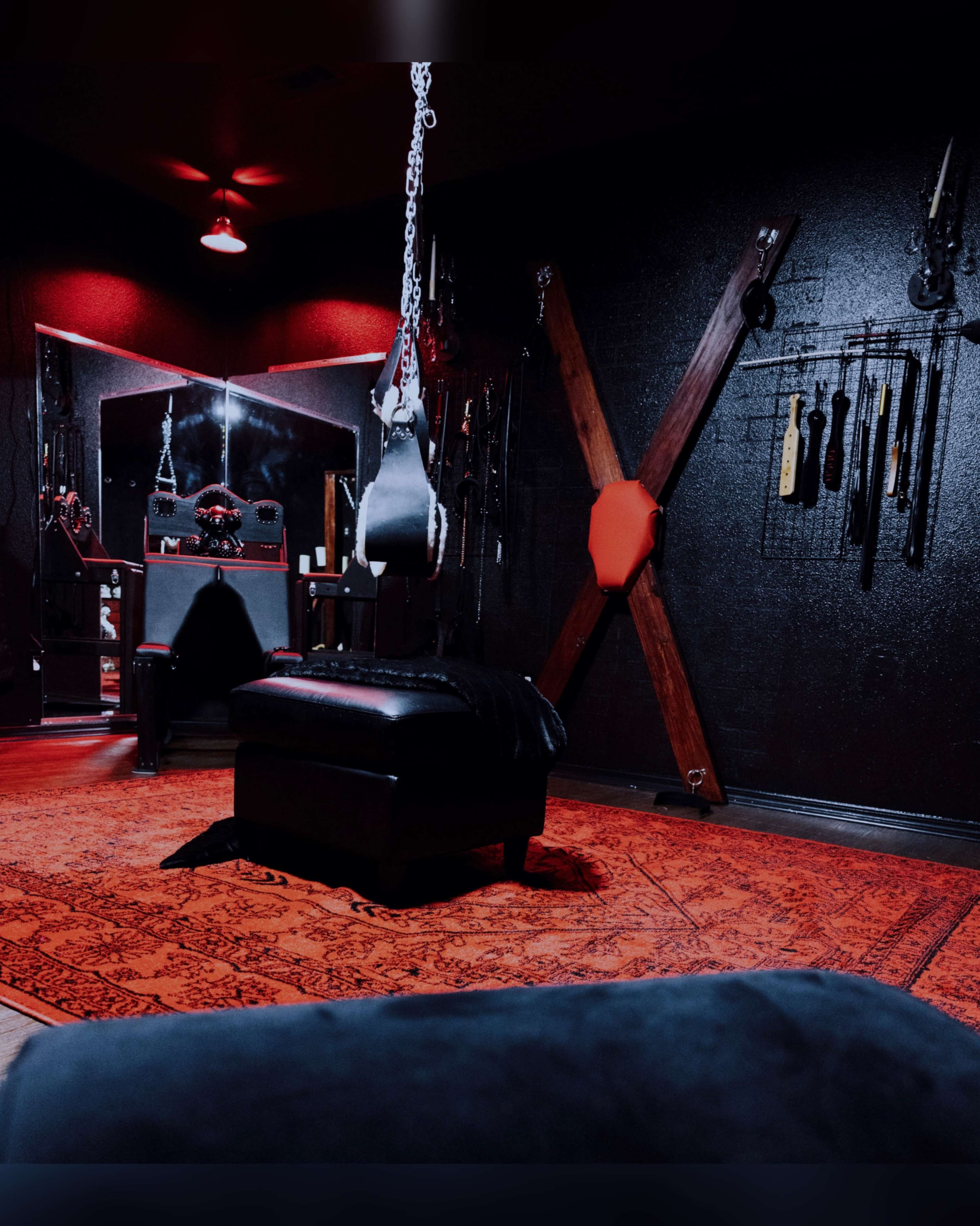 The room features a dark interior with red lighting, a mirror, a black padded bench, and various tools hanging on the walls.
