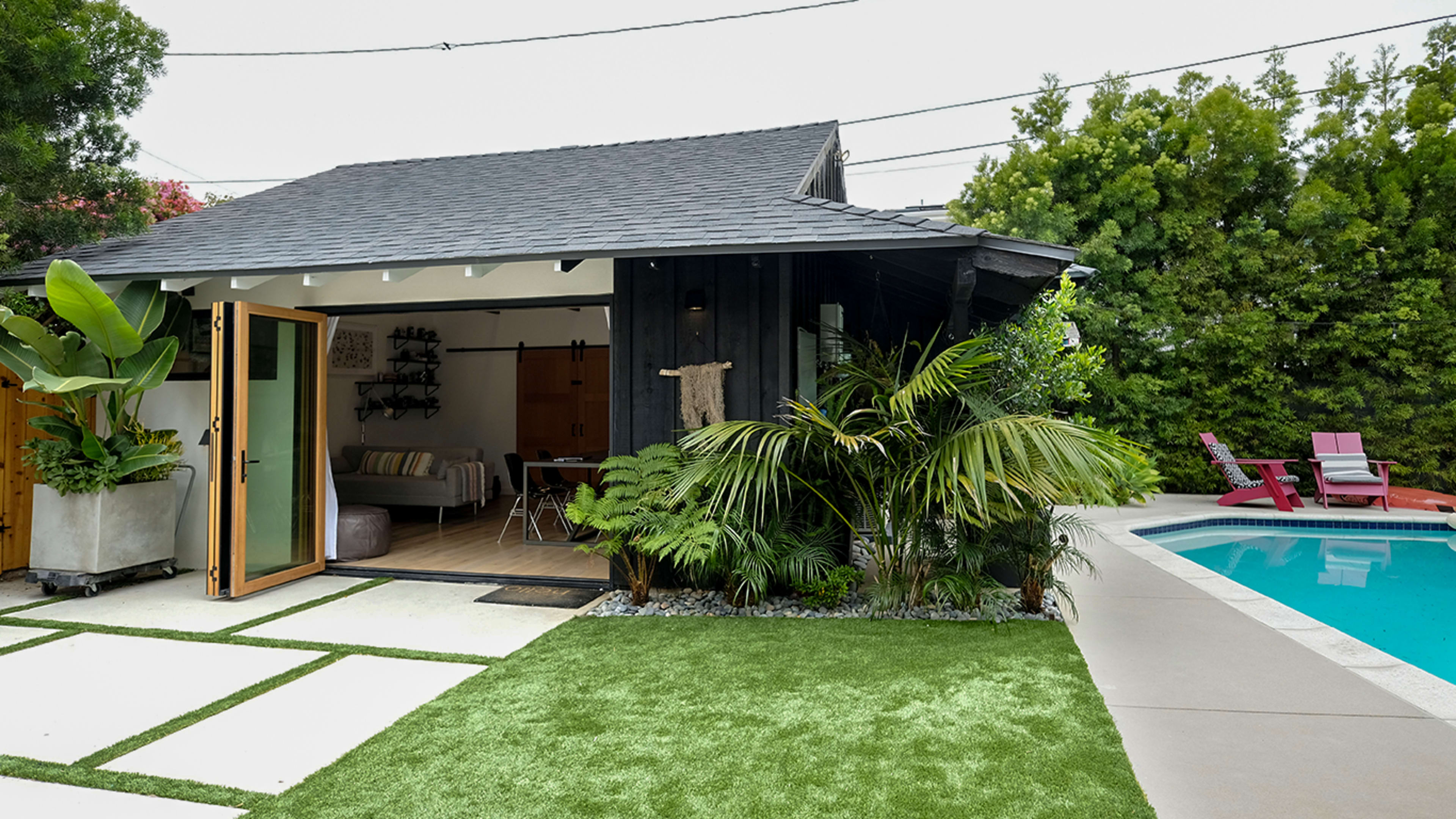 A small modern building with large sliding doors is surrounded by greenery and features a swimming pool nearby.