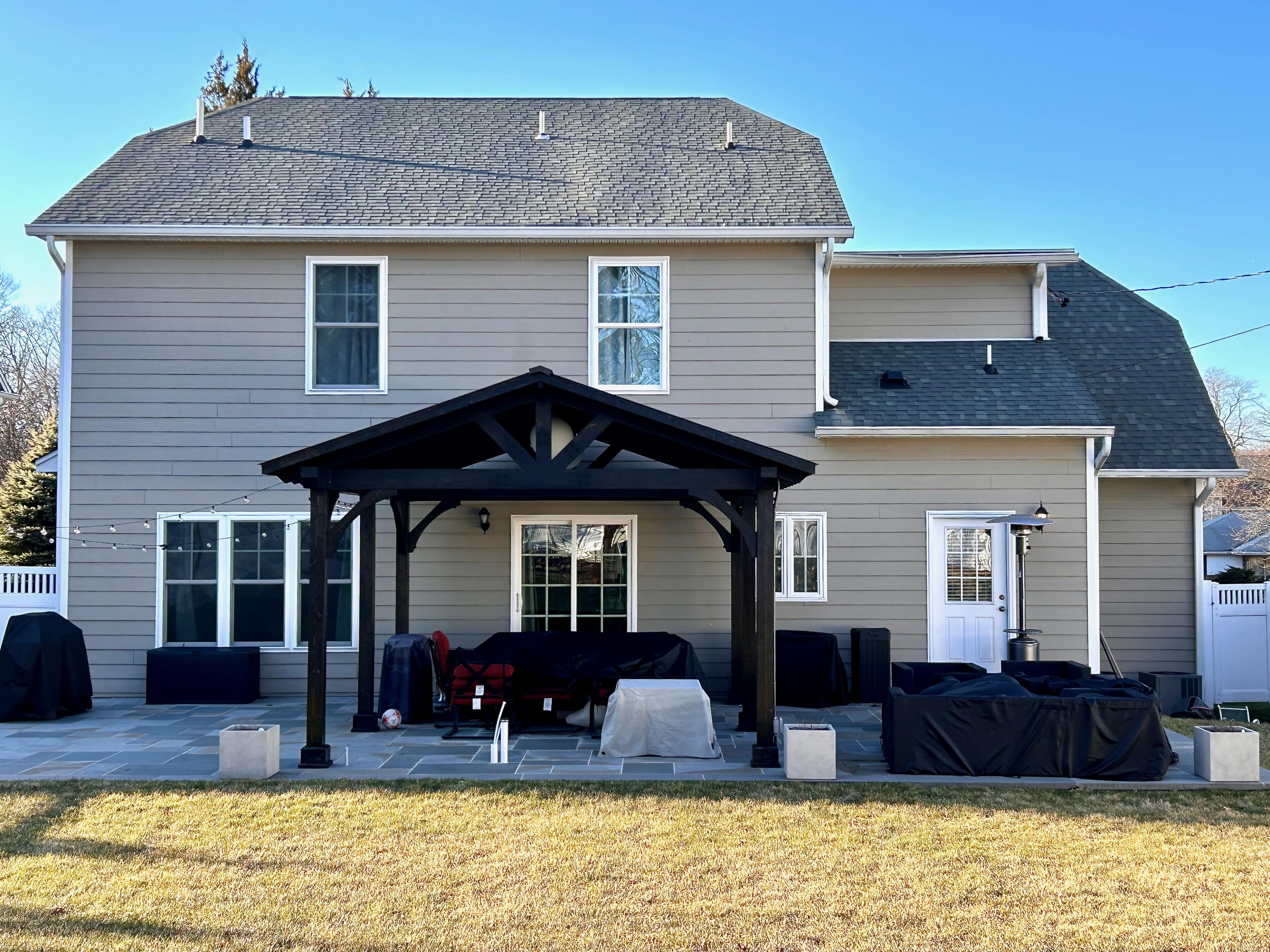 A two-story house with a covered patio, surrounded by a grassy yard and several outdoor furniture pieces covered with protective tarps.