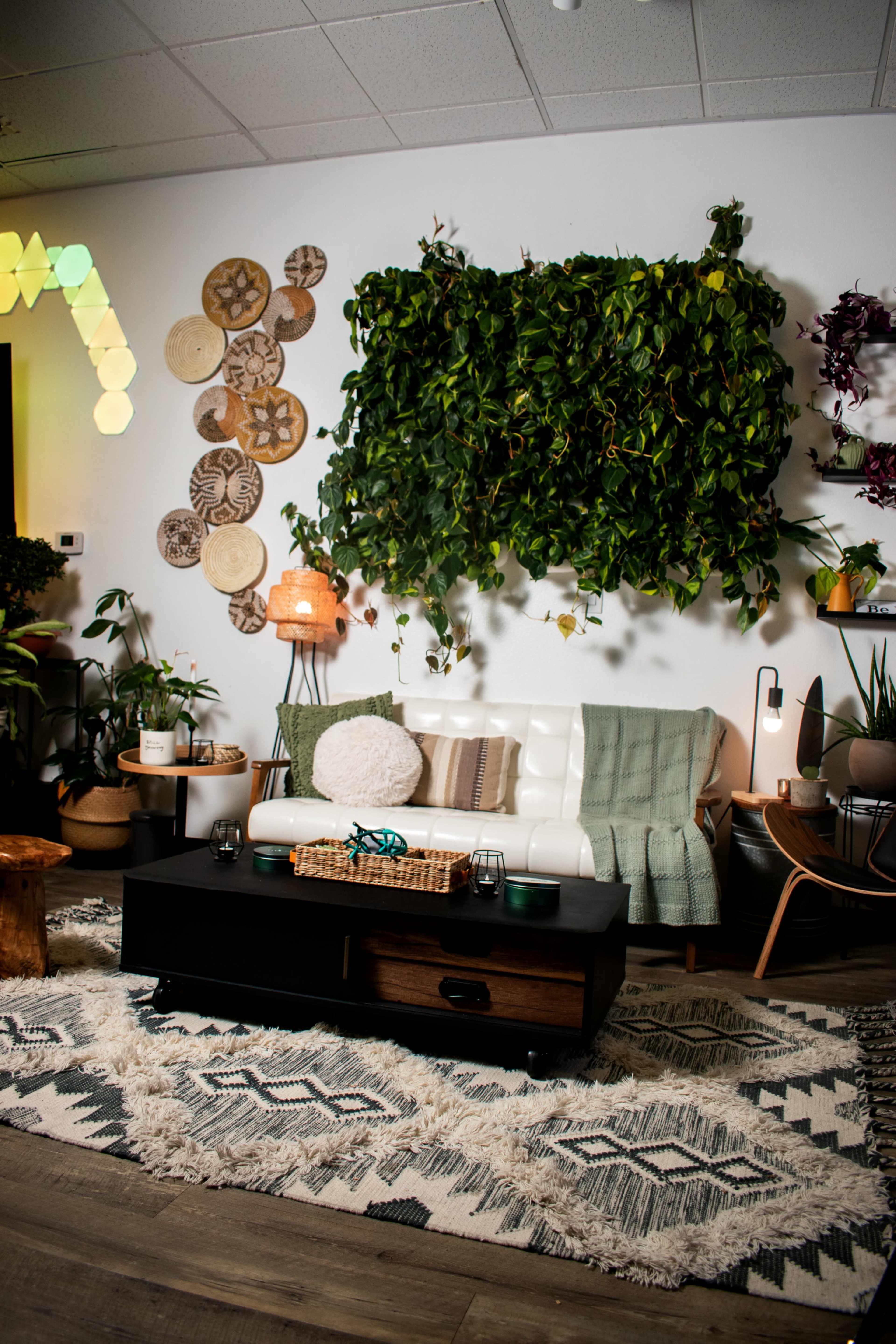 A modern living room features a white sofa, a coffee table, various plants, wall art made from natural materials, and a textured area rug.