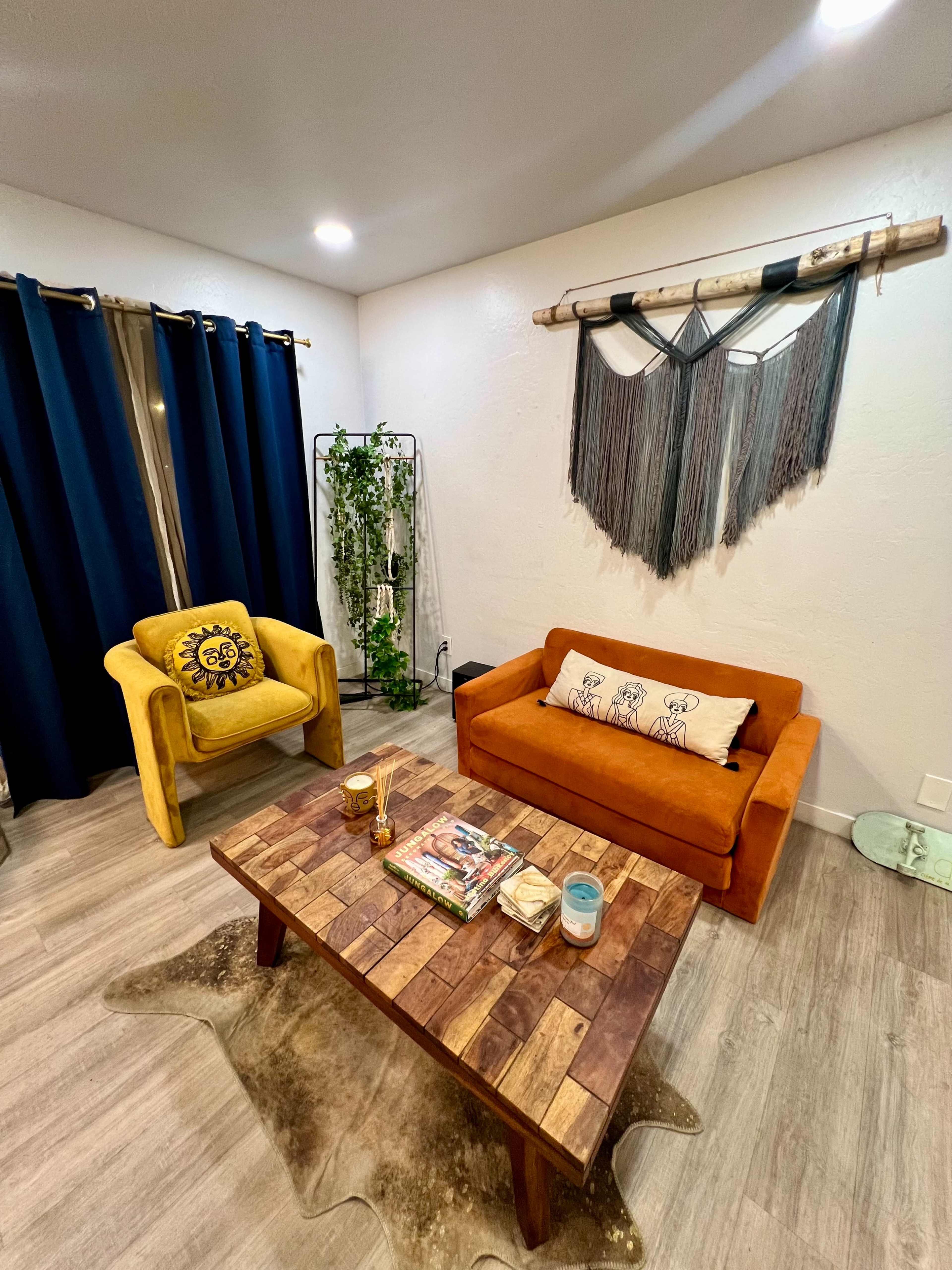 A cozy living room with two chairs, a wooden coffee table, and wall decor featuring a textile hanging, set against a backdrop of dark curtains and a potted plant.