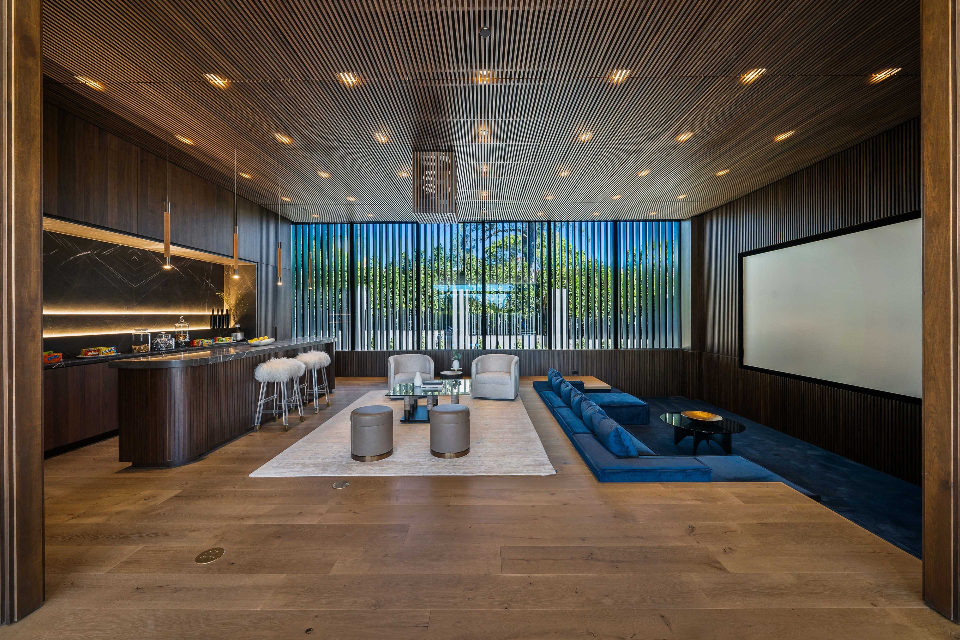 The image shows a modern living space with a sleek bar area, a large screen for viewing, and seating arrangements featuring a blue sofa and round stools on a light wooden floor.