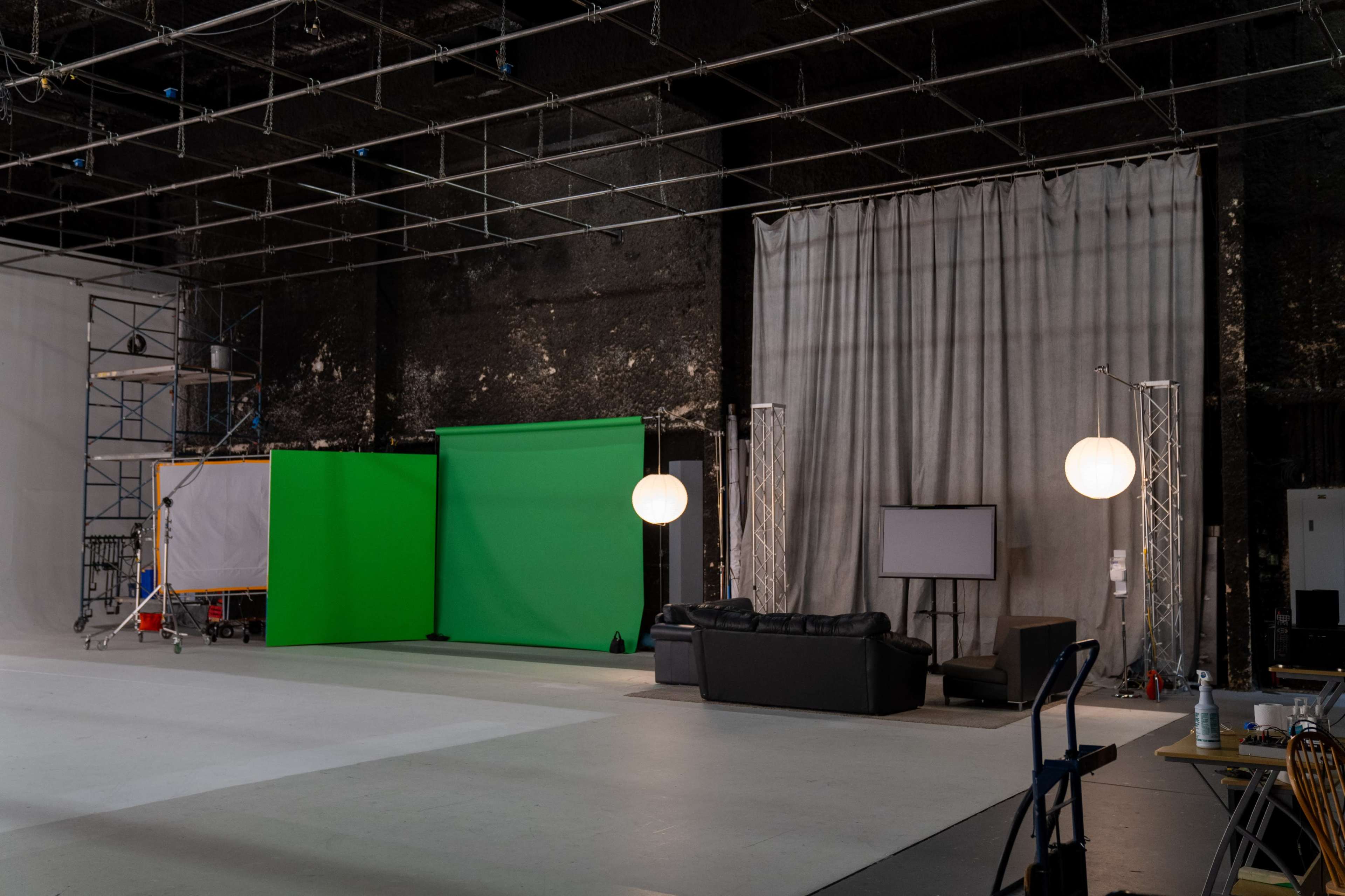 A film studio setup with a green screen, a gray backdrop, a black couch, a television on a stand, and soft lighting fixtures.