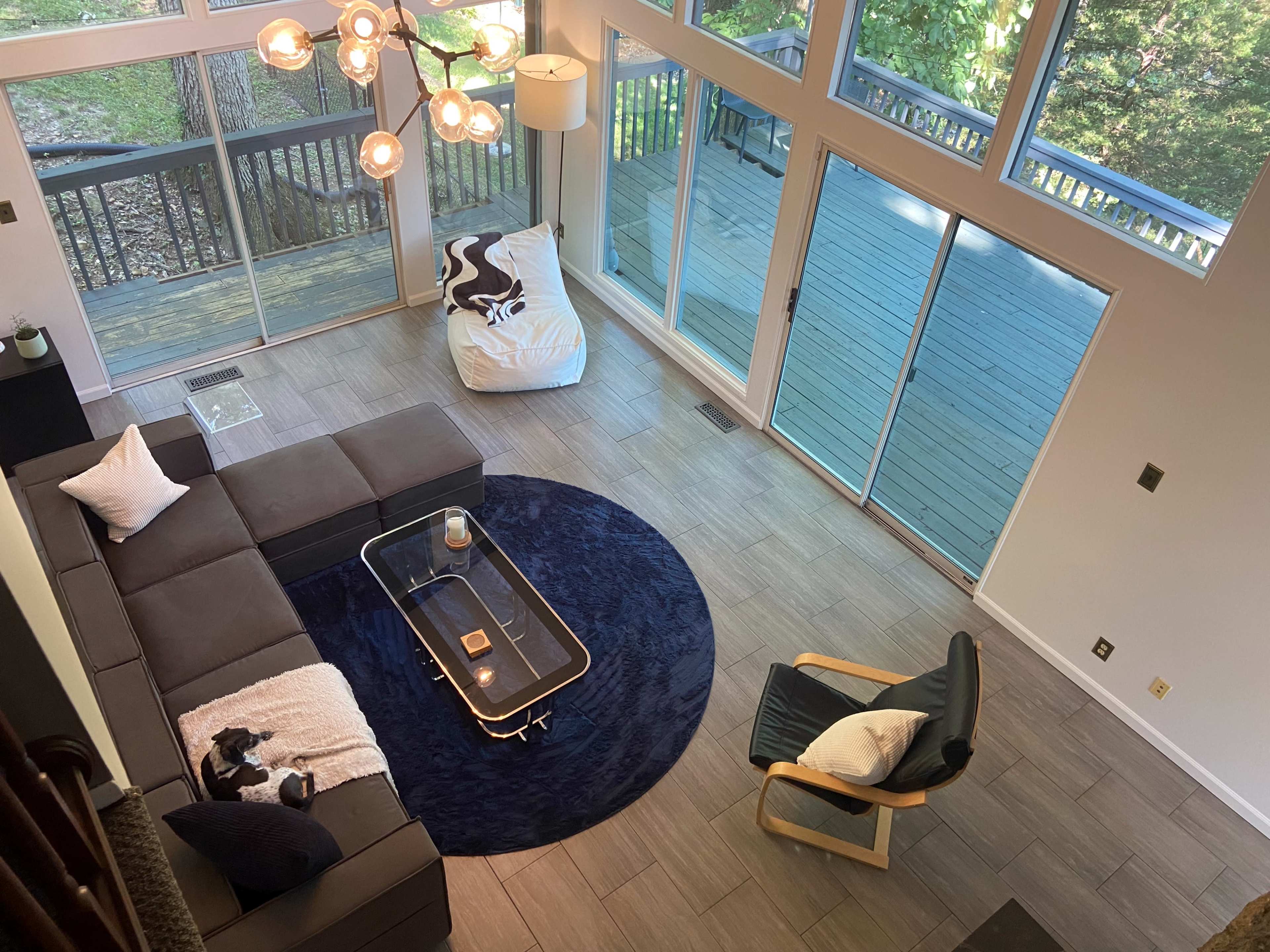 A modern living room features a sectional sofa, a round rug, a glass coffee table, and large windows overlooking a deck with trees in the background.