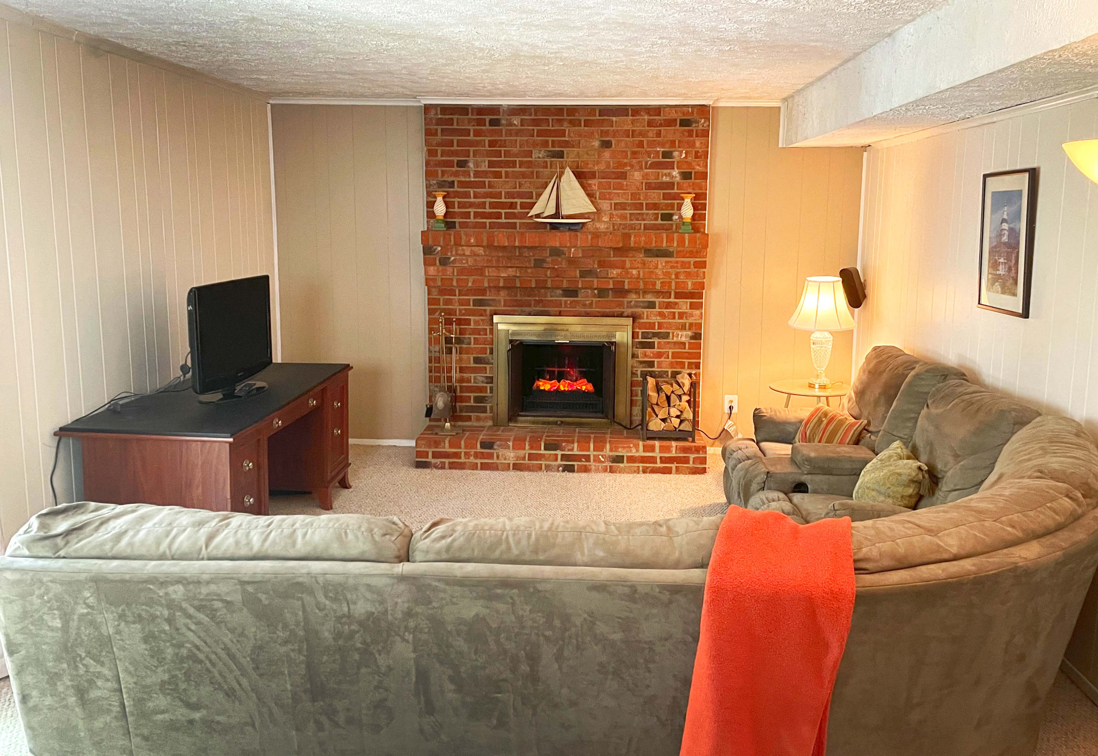 A cozy living room features a large sectional sofa, a brick fireplace with a decorative sailboat, a television on a wooden stand, and a lamp beside the couch.