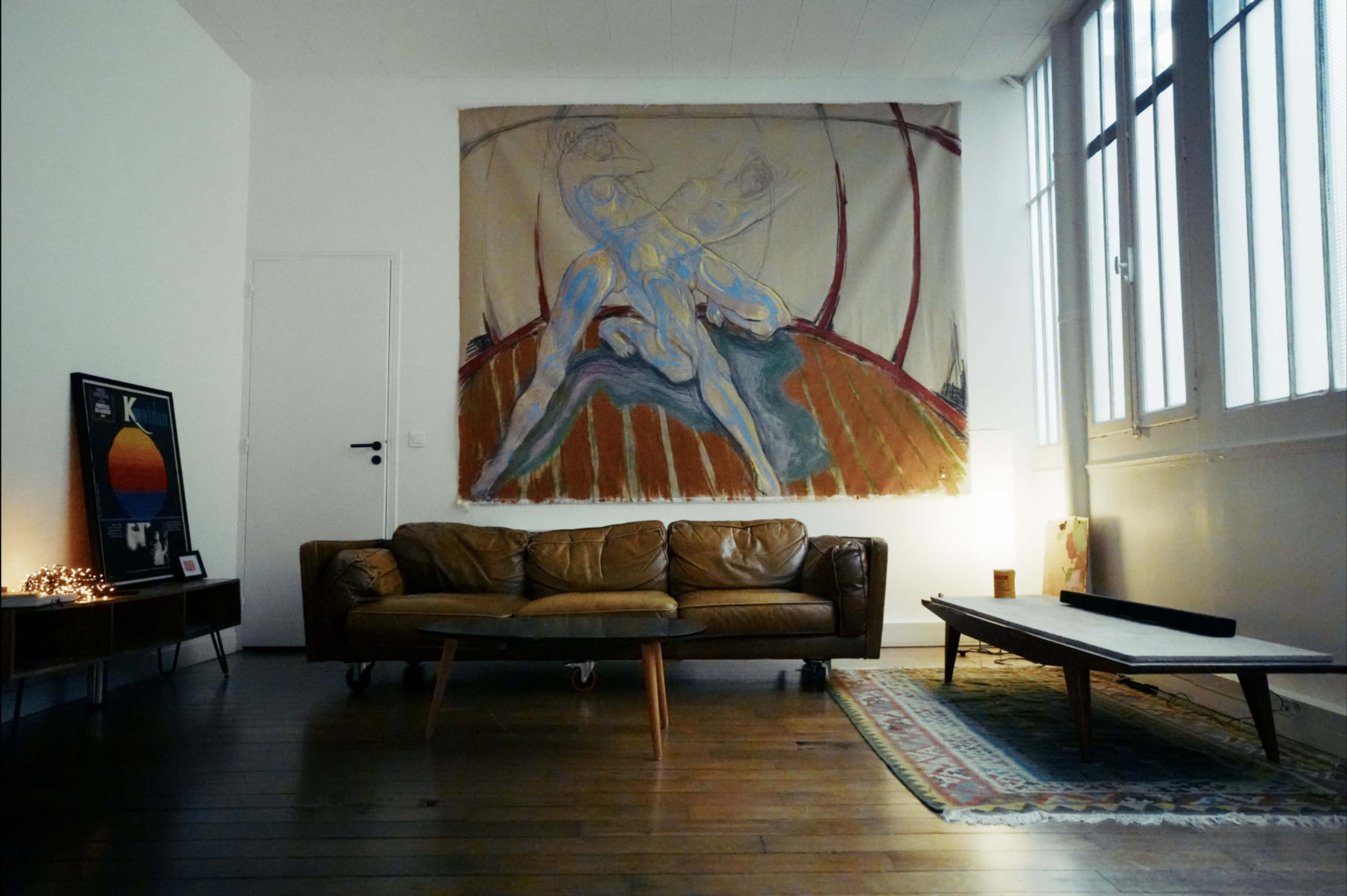 The image shows a living room with a large artwork on the wall, a brown leather sofa in the center, and a wooden coffee table on a patterned rug.