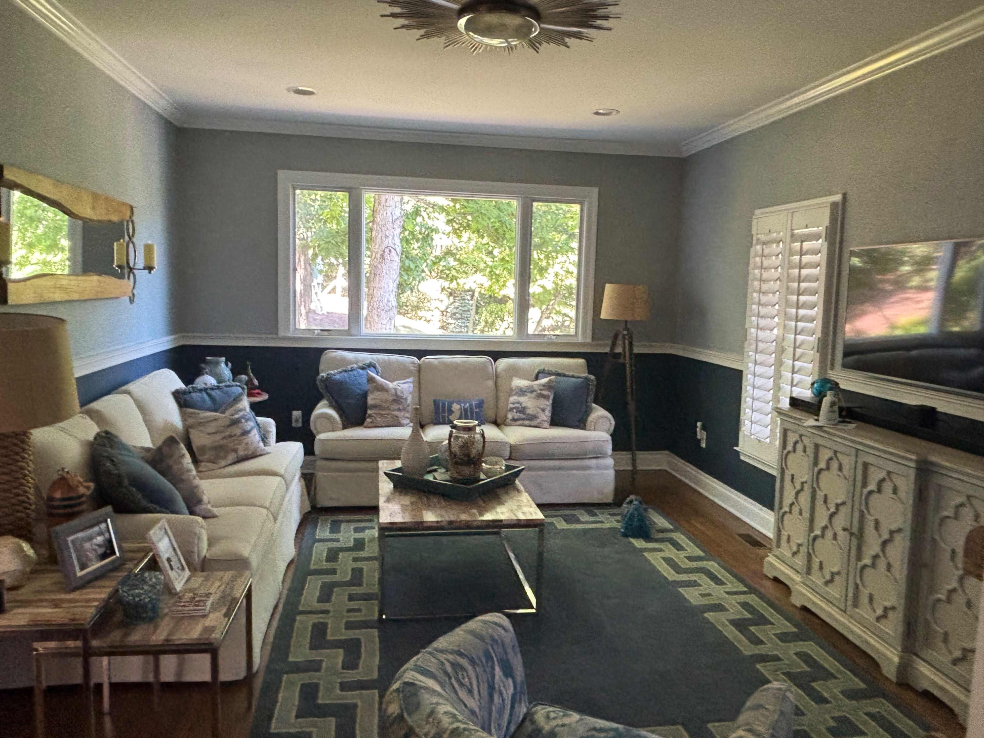 The living room features two white sofas, a coffee table with decorative items, and large windows that allow natural light to fill the space.